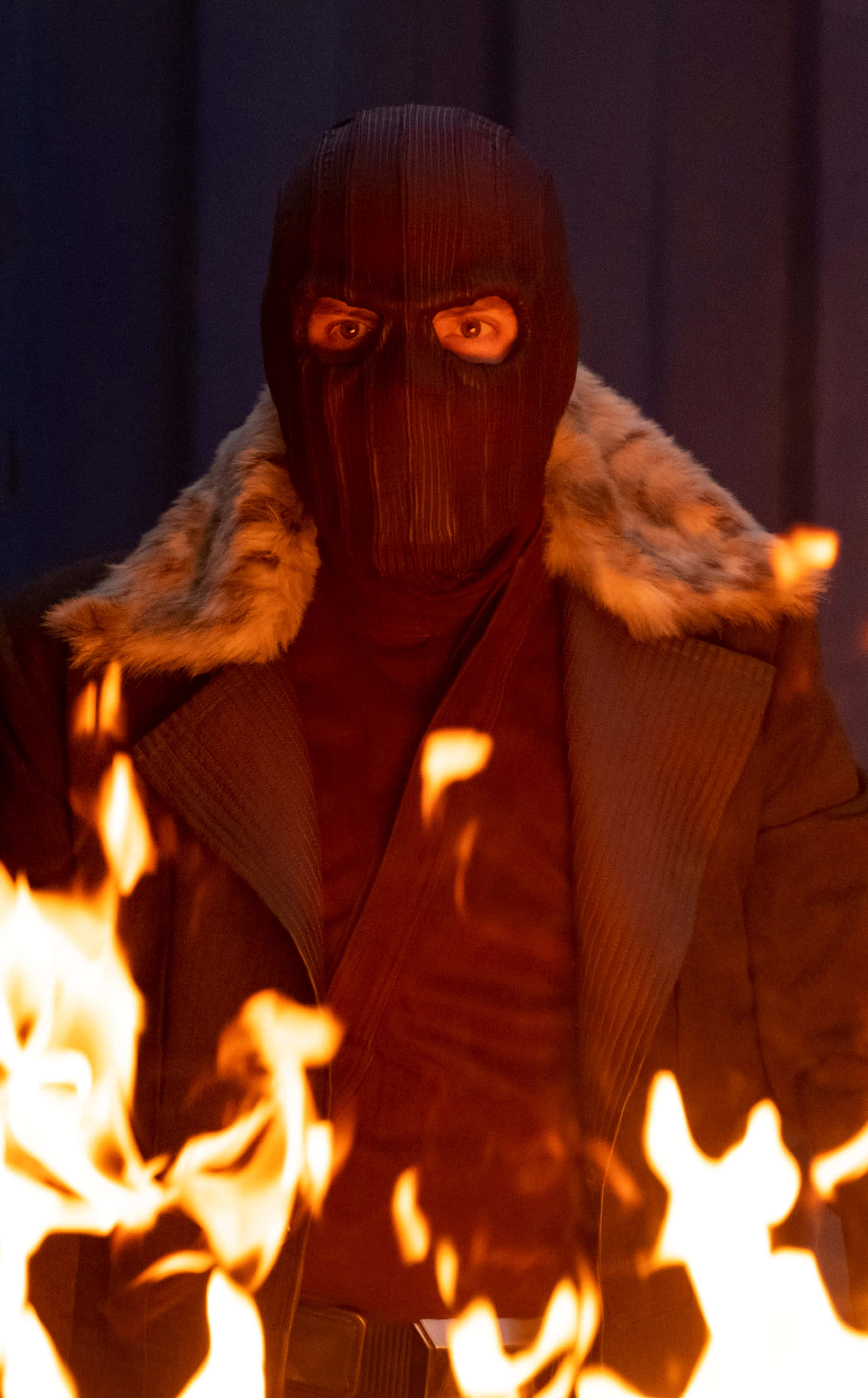 Baron Zemo 桌布 壁紙 — 一個戴著面具、身穿毛皮襯裡大衣的人物站在咆哮的火焰後面，臉上投射出強烈的橙色光芒。