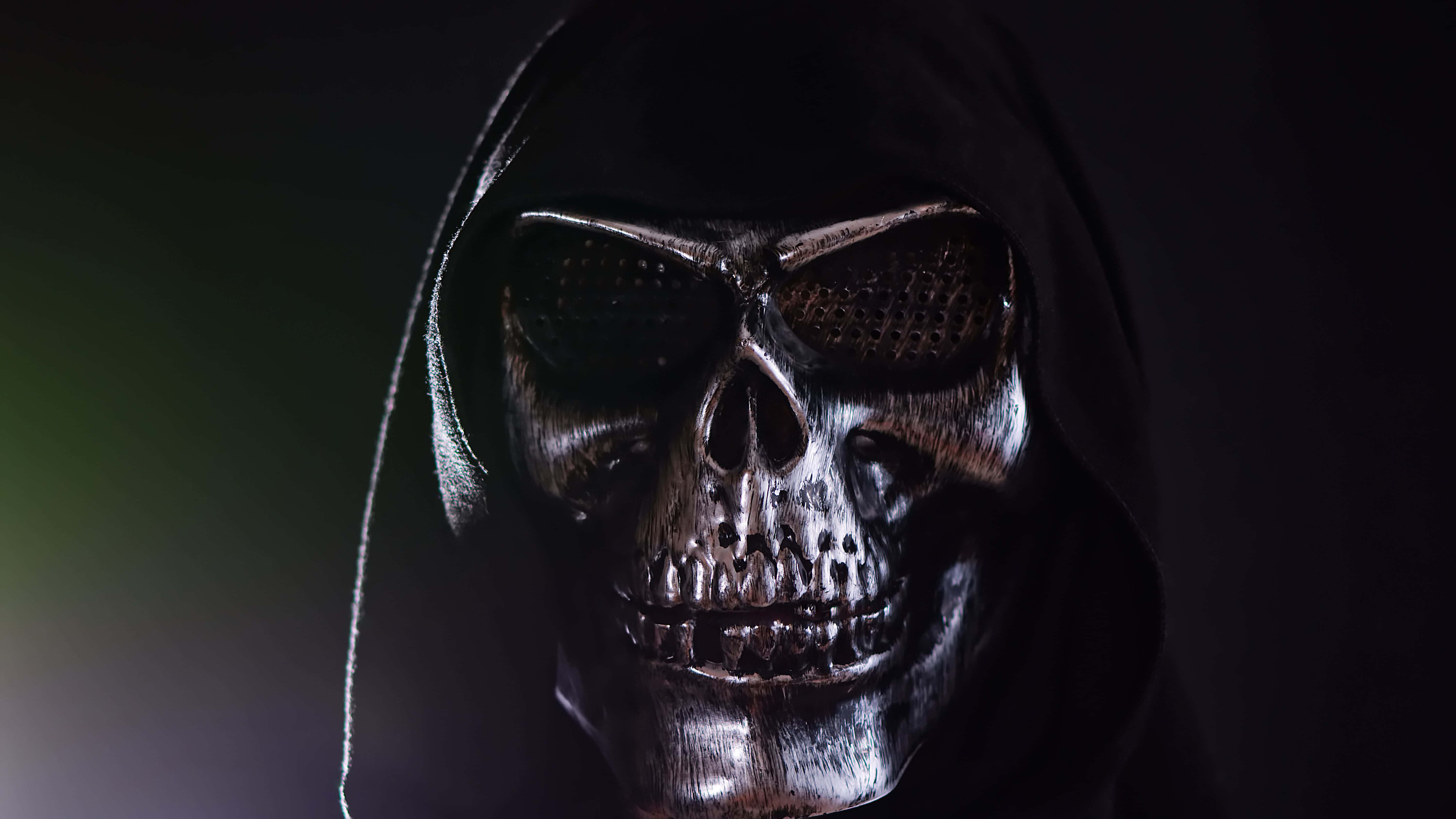 Halloween Masks 桌布 壁紙 — 一個戴著頭罩、帶有網狀眼罩的威脅性骷髏頭，在黑暗背景下於戲劇性燈光下發光。