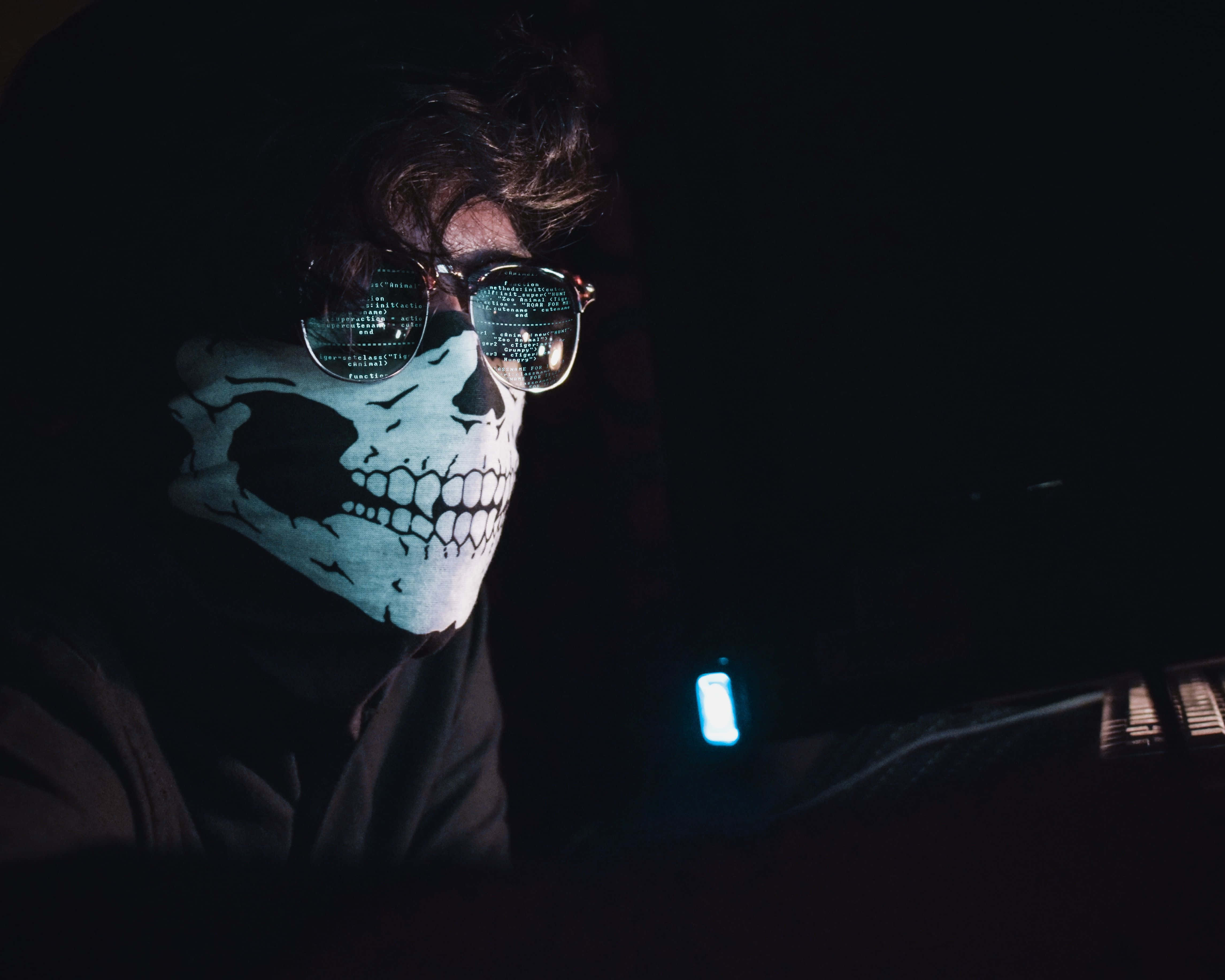 Horror Masks 桌布 壁紙 — 一個戴著骷髏面具和眼鏡的人，眼鏡反射著數碼代碼，坐在黑暗中，被發光的藍色屏幕照亮。