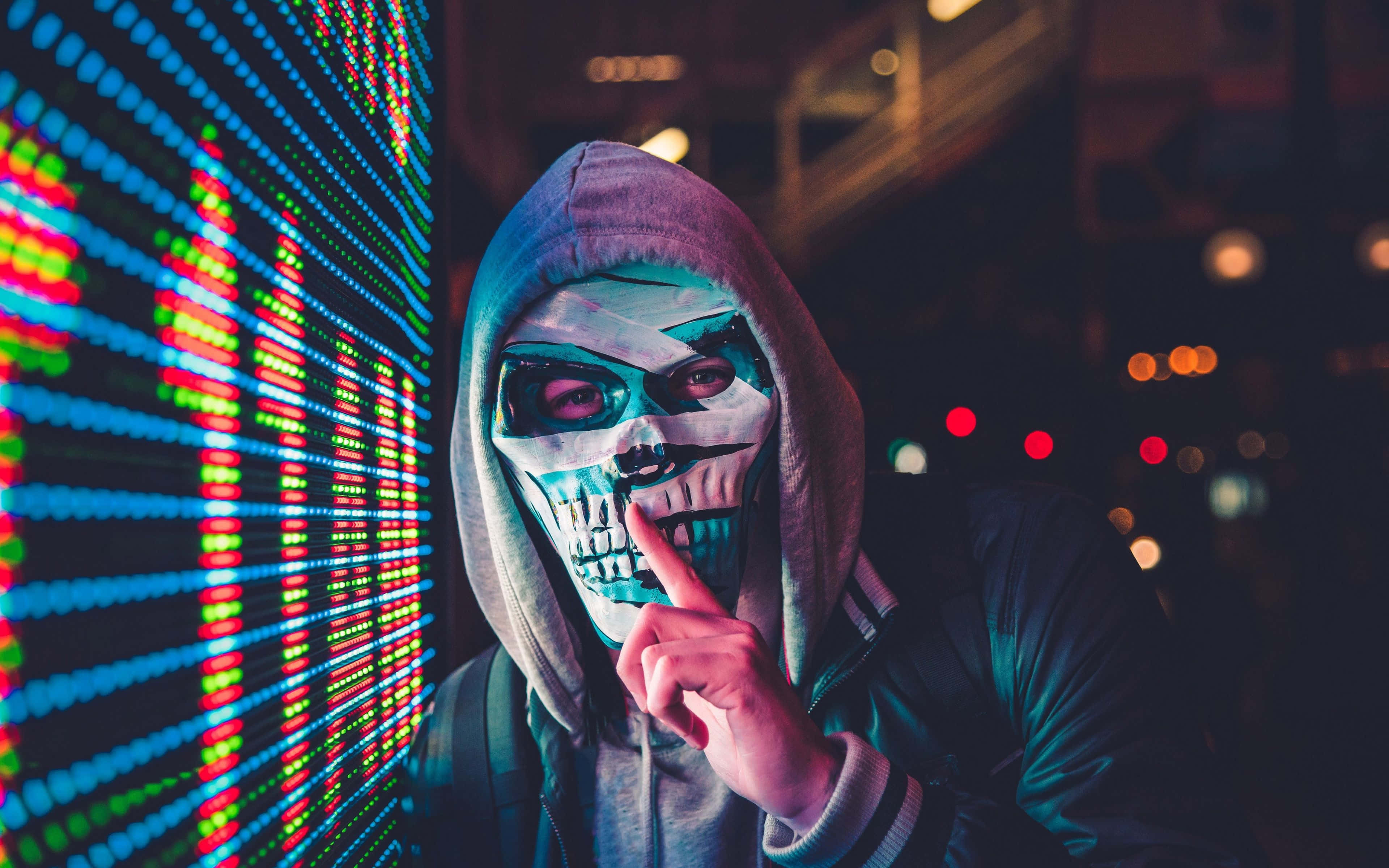 Horror Masks 桌布 壁紙 — 一個戴著骷髏面具和連帽衫的人站在黑暗城市夜晚環境中發光的數碼屏幕前。