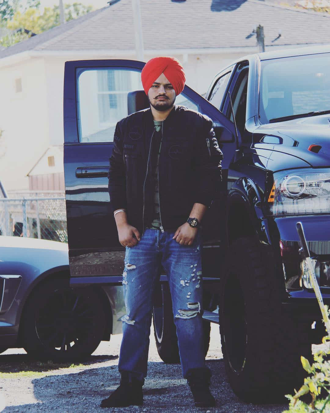 Sidhu Moose Wala 桌布 壁紙 — 一個戴著鮮紅色頭巾的男人在陽光明媚的日子裡自信地站在一輛打開車門的黑色皮卡車旁。
