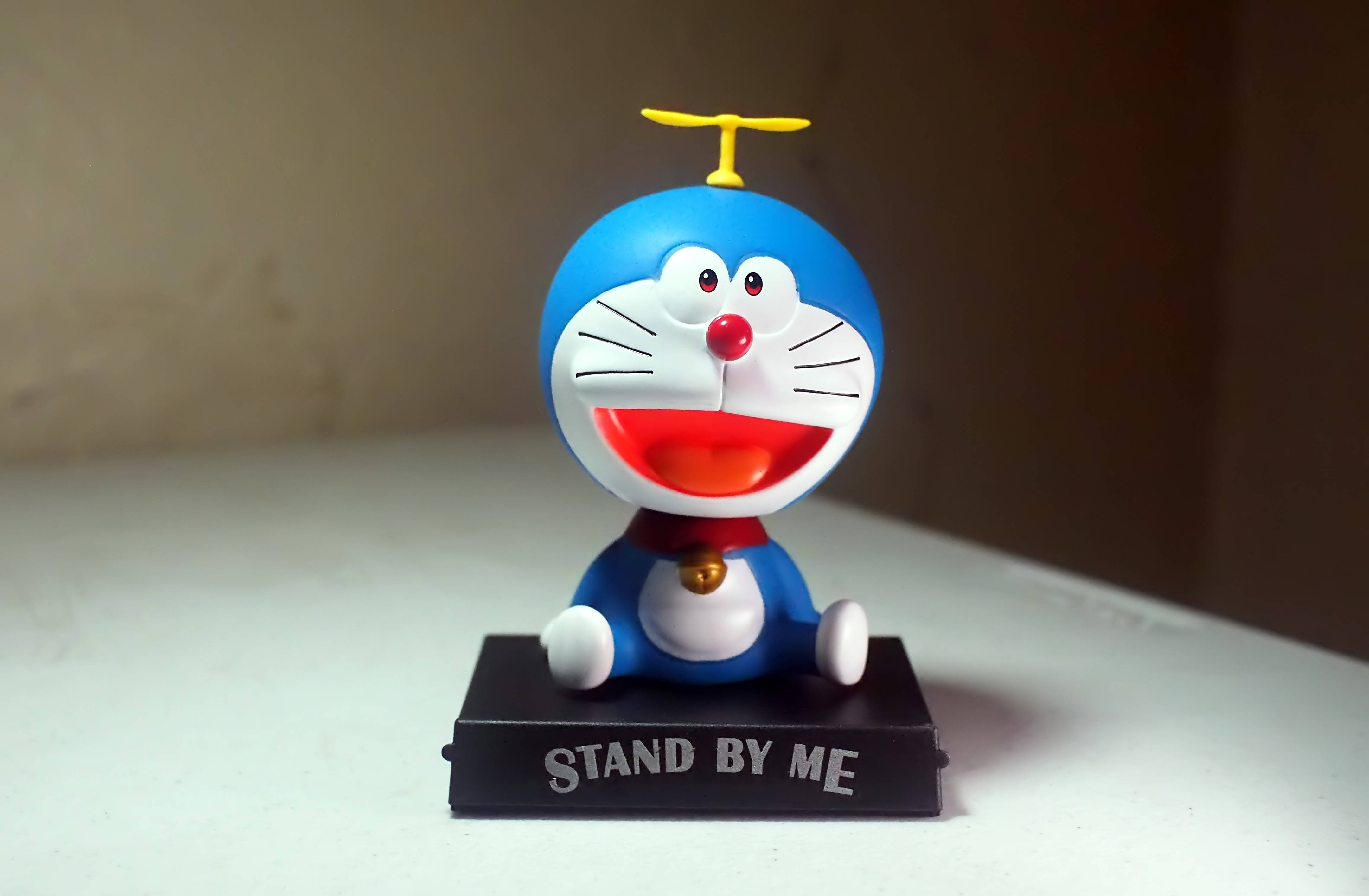 Doraemon 桌布 壁紙 — 一個戴著黃色螺旋槳帽的開心藍色機械貓公仔，坐在黑色底座上，底座上寫著「STAND BY ME」，放在白色表面上。