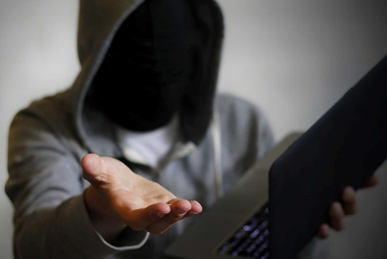 Blackmail 桌布 壁紙 — 一個戴著黑色面具的兜帽人物，在昏暗的房間裡拿著手提電腦，同時向觀看者伸出手。