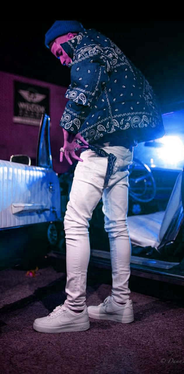 NLE Choppa 桌布 壁紙 — 一個戴頭巾外套、穿白褲的人，在霓虹藍光下靠在一輛復古車旁。
