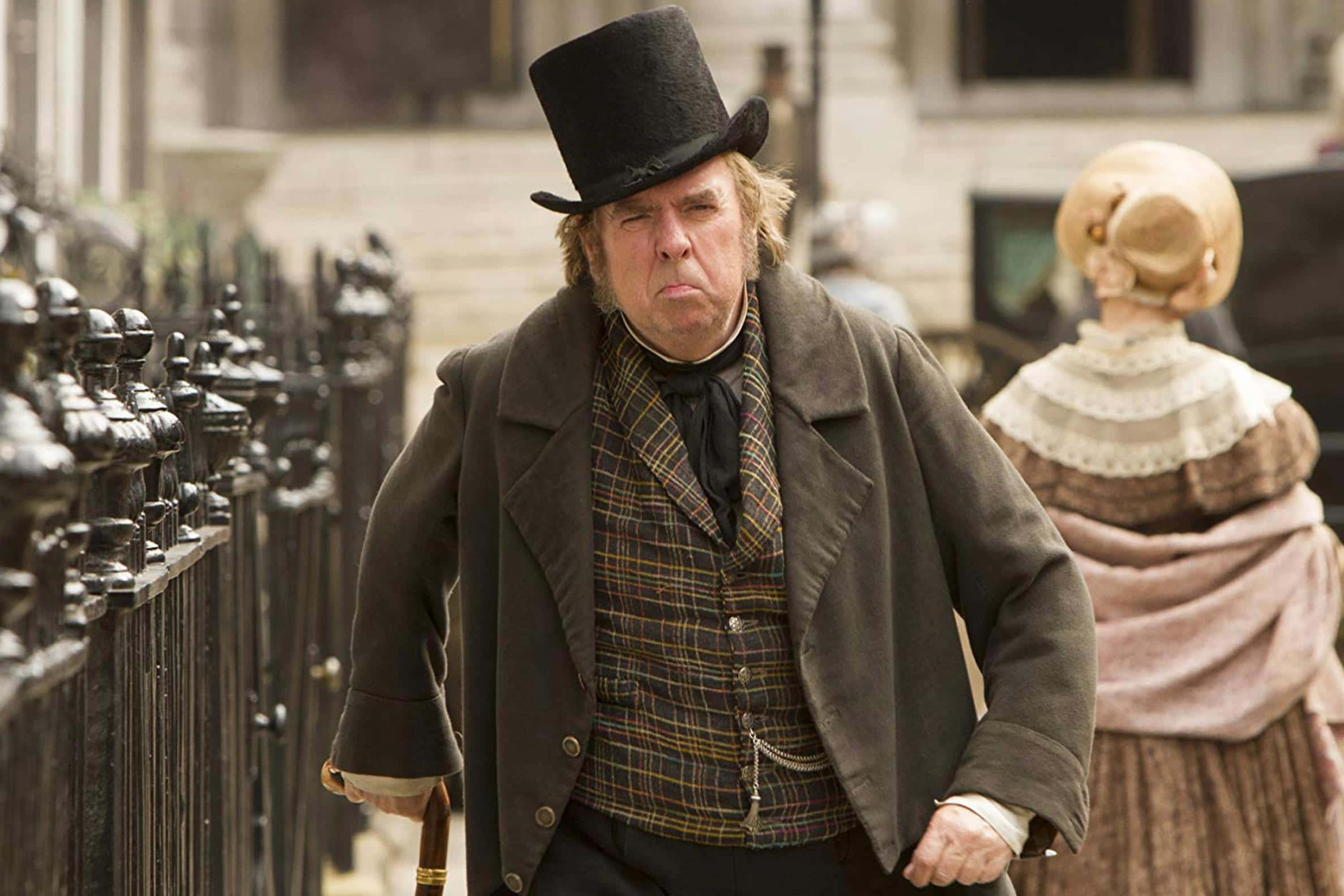 Timothy Spall 桌布 壁紙 — 一個戴高帽、穿格仔背心的暴躁男人，身後跟着一個戴帽子的女人，走在鵝卵石街上。
