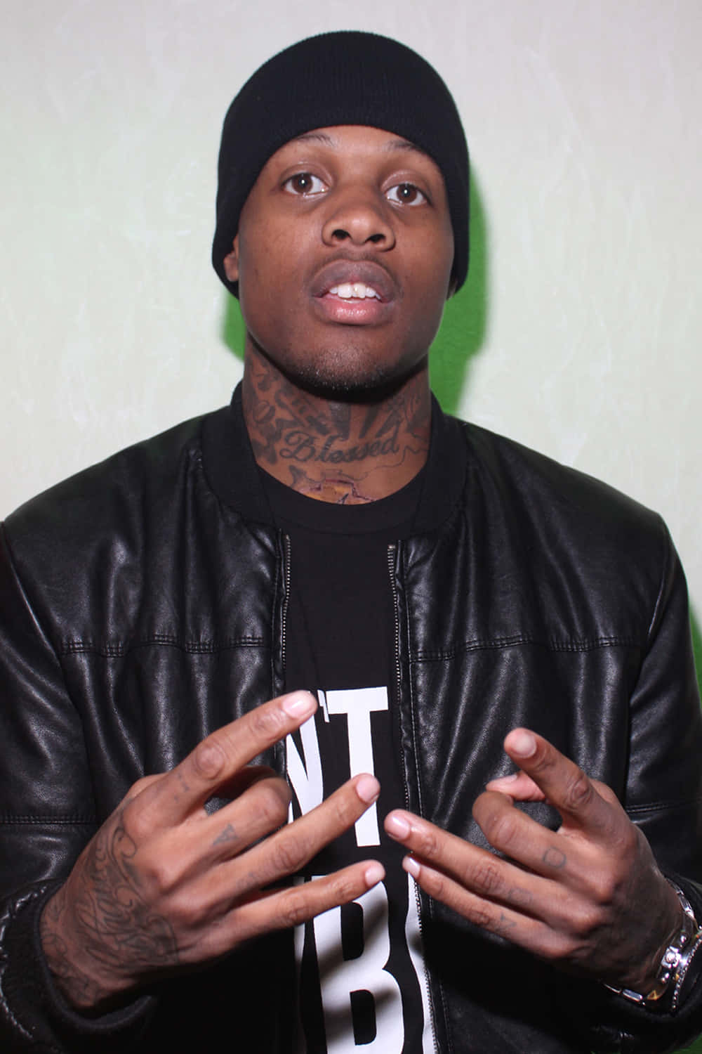 lil durk 桌布 壁紙 — 一個戴黑色冷帽、穿皮褸的男人，在純綠色牆前說話時做手勢。