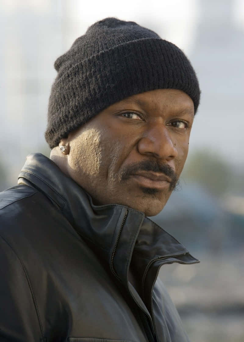 Ving Rhames 桌布 壁紙 — 一個戴黑色冷帽和皮褸的男人，在模糊的城市背景下，神情嚴肅地凝視着。