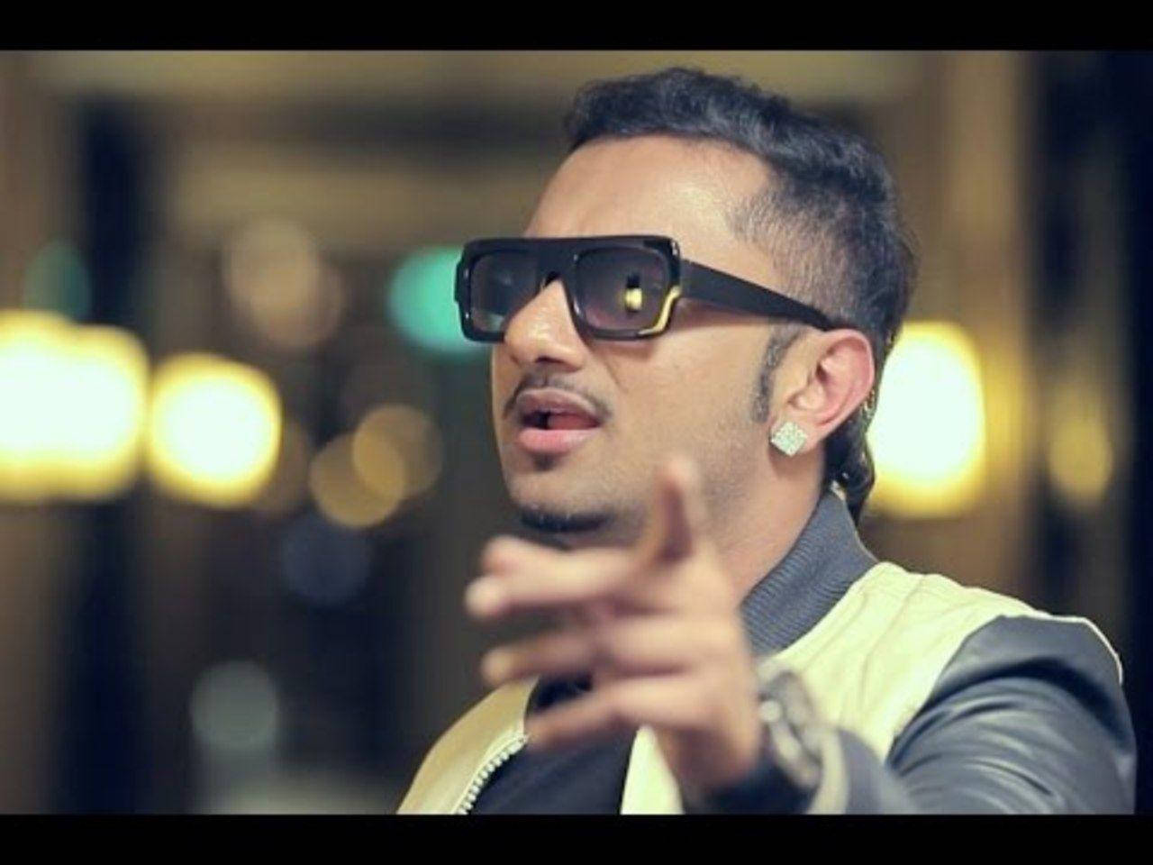 Yo Yo Honey Singh Sam Sandhu 壁紙 — 一個戴黑色太陽眼鏡和皮褸的男人，在模糊的金色散景背景前指向前面。