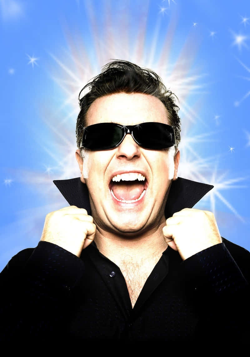 ricky gervais 桌布 壁紙 — 一個戴黑色太陽眼鏡和穿黑色襯衫的男人，在明亮的藍色星爆背景下勝利地大喊。