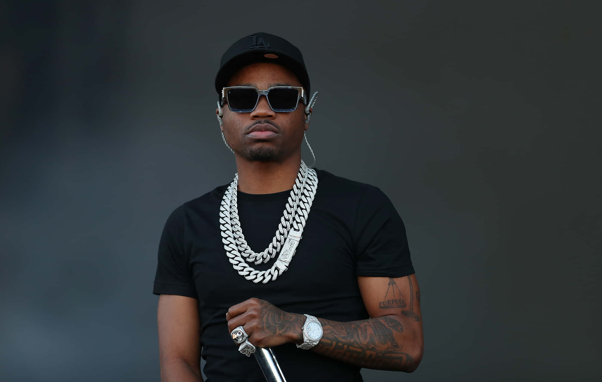 Roddy Ricch 桌布 壁紙 — 一個戴黑色帽和太陽眼鏡的男人，戴著一條粗銀鏈，在深灰色背景前拿著咪高峰。