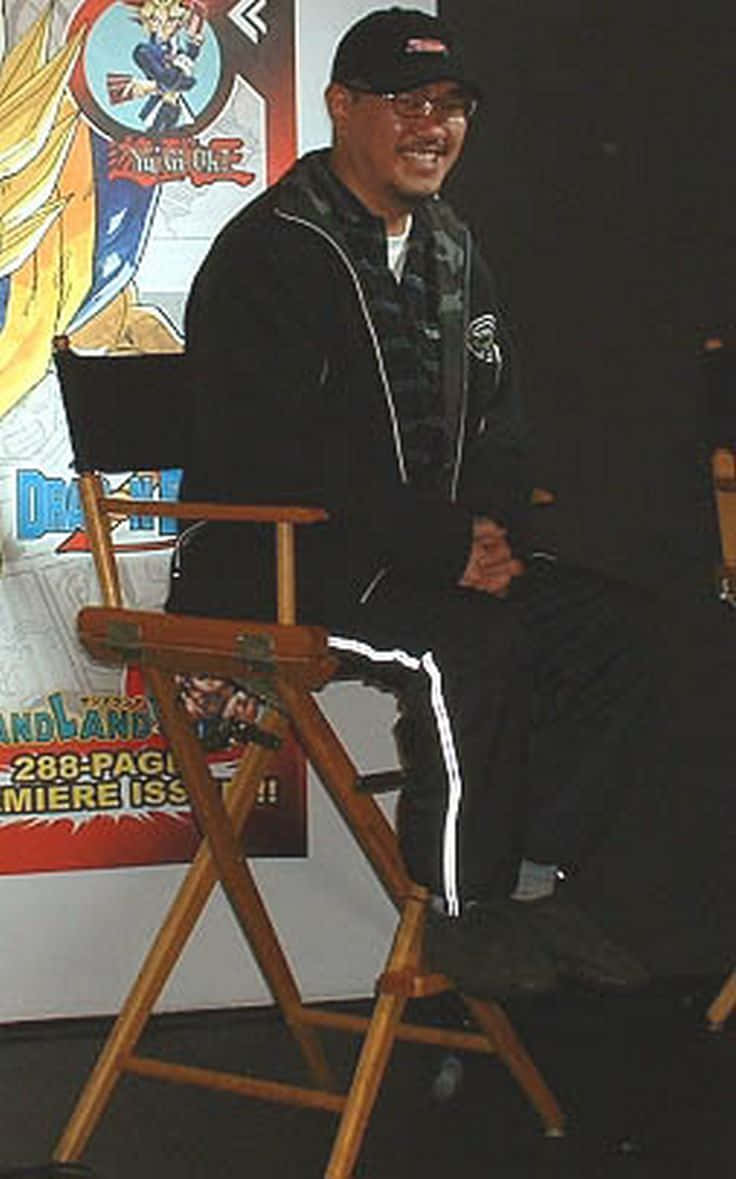 Akira Toriyama 桌布 壁紙 — 一個戴黑色帽和穿黑色外套的微笑男人坐在木製導演椅上，前面有一張龍珠海報。