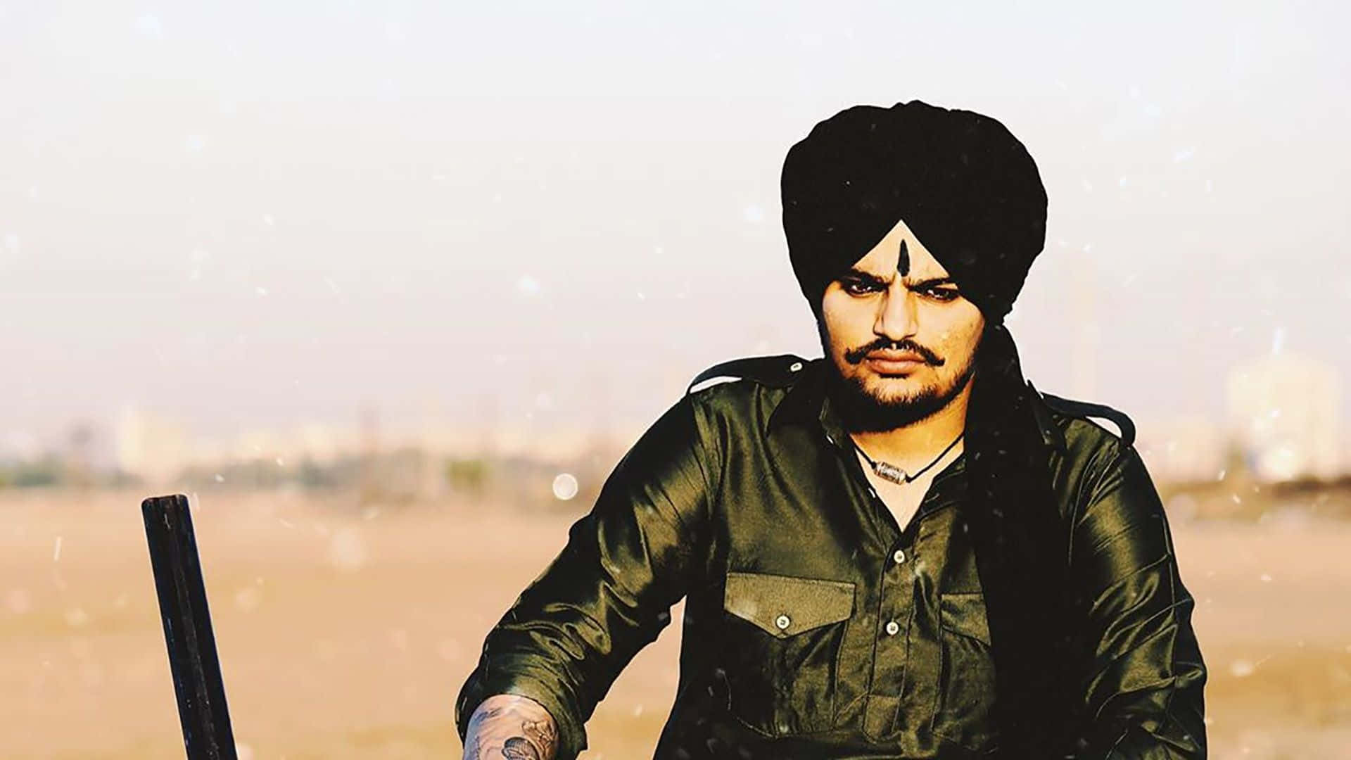 Sidhu Moose Wala 桌布 壁紙 — 一個戴黑色頭巾、穿綠色襯衫的男人自信地站在一個模糊的城市天際線背景下的朦朧戶外環境中。
