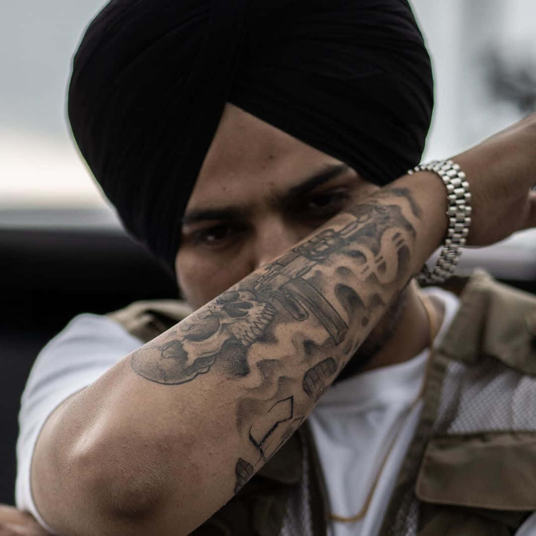 Sidhu Moose Wala 桌布 壁紙 — 一個戴黑色頭巾的男人用他紋有骷髏和槍的手臂遮住臉。