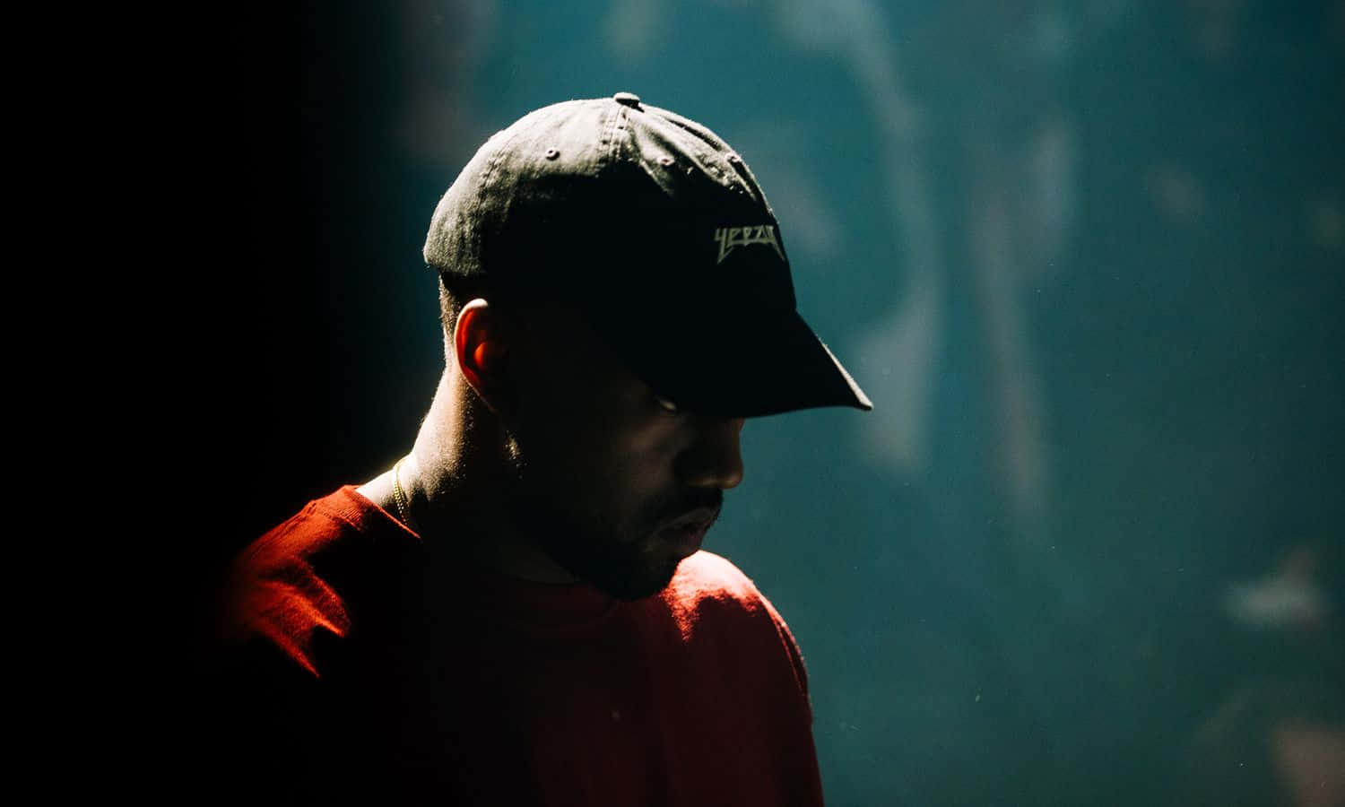 Kanye 桌布 壁紙 — 一個戴黑色鴨舌帽、穿紅色襯衫的男人，在朦朧的藍綠色背景上，以戲劇性的側光呈現剪影。