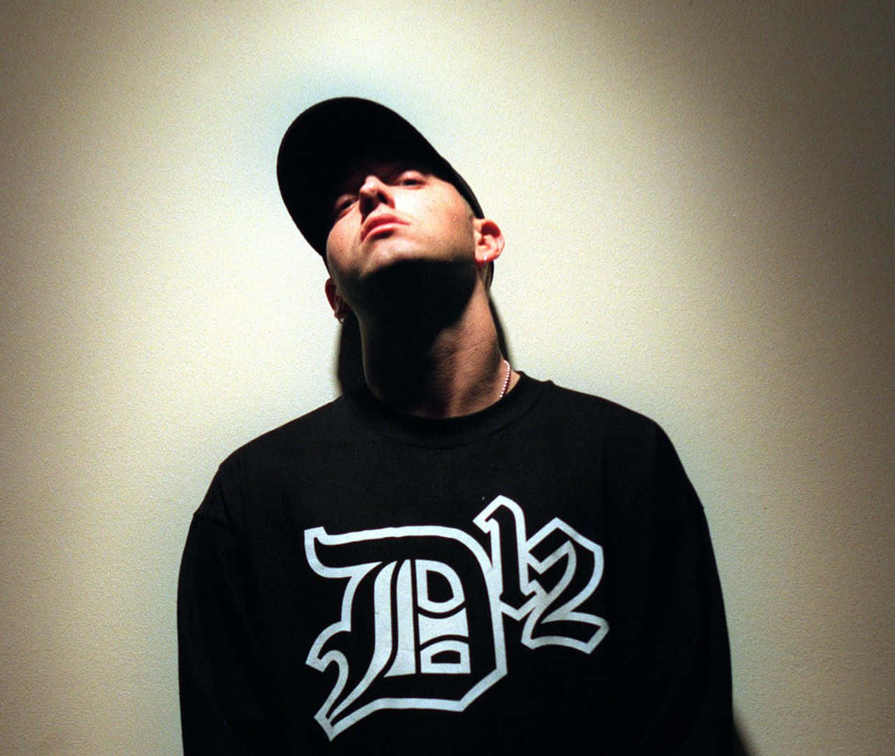 Eminem 桌布 壁紙 — 一個戴黑色鴨舌帽、身穿D12衛衣嘅男人，喺戲劇性嘅射燈下對住一幅素牆向上望。