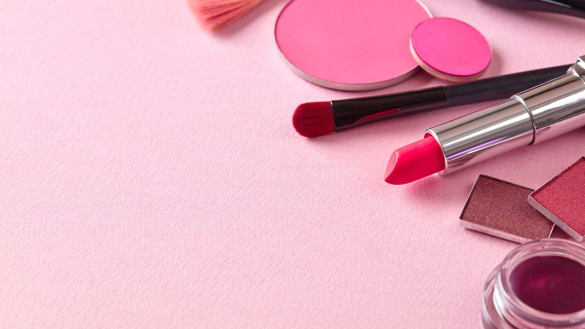 Pink Makeup 桌布 壁紙 — 一個扁平擺放的鮮豔粉紅色和紅色化妝品，包括唇膏、刷子、眼影和唇彩，排列在柔軟的粉紅色紋理表面上。
