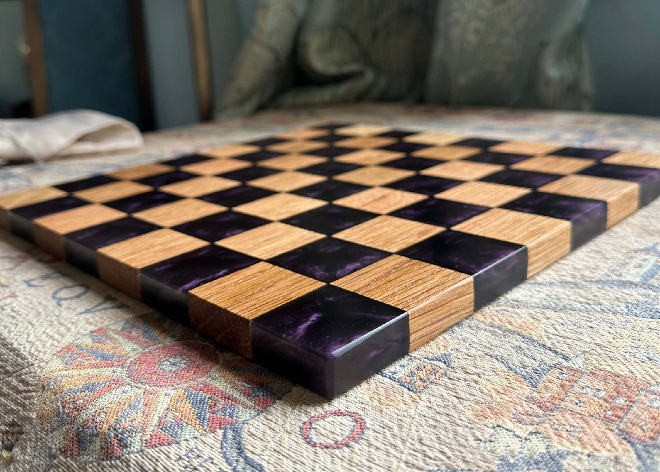 Chessboard 桌布 壁紙 — 一個手工製作的木製國際象棋棋盤，帶有紫色樹脂方格，放置在帶有圖案的紡織品表面上。