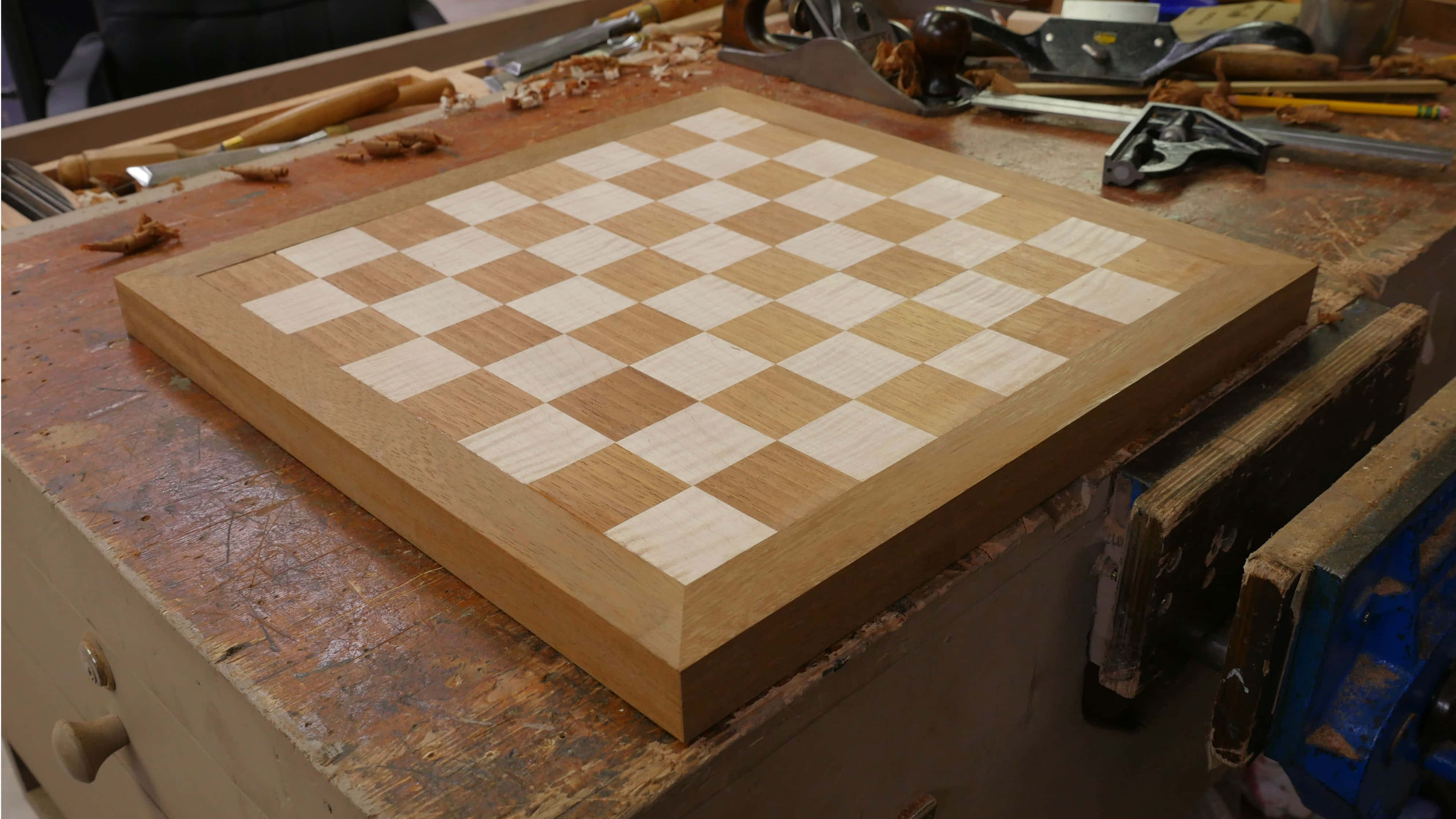Chessboard 桌布 壁紙 — 一個手工製作的木製國際象棋棋盤，帶有淺色和深色橡木方格，放置在雜亂的木匠工作台上，周圍環繞著工具。