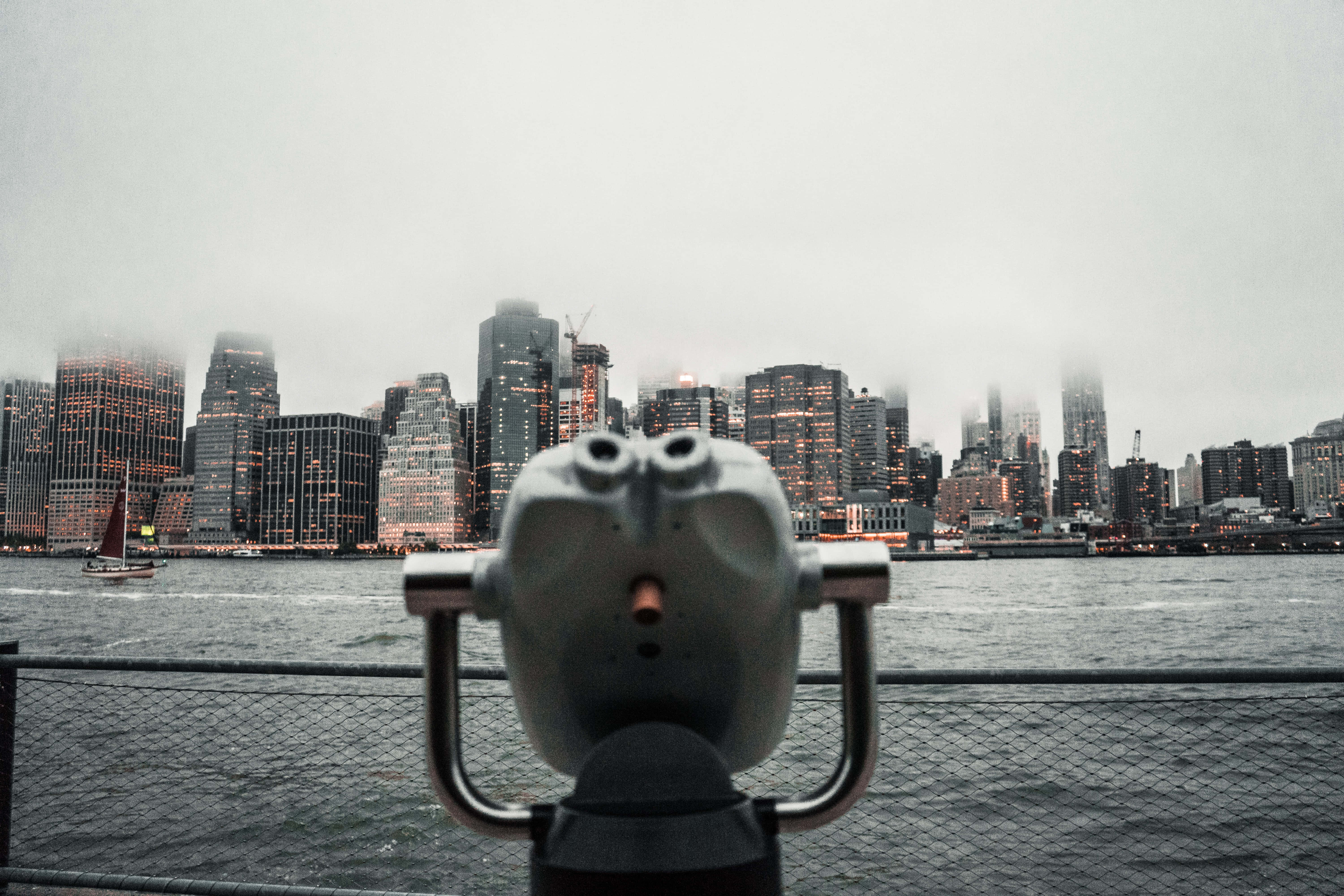 Nyc 桌布 壁紙 — 一個投幣式雙筒望遠鏡框住了霧氣瀰漫的紐約市天際線，窗戶閃爍著光芒，水面上有一艘帆船。