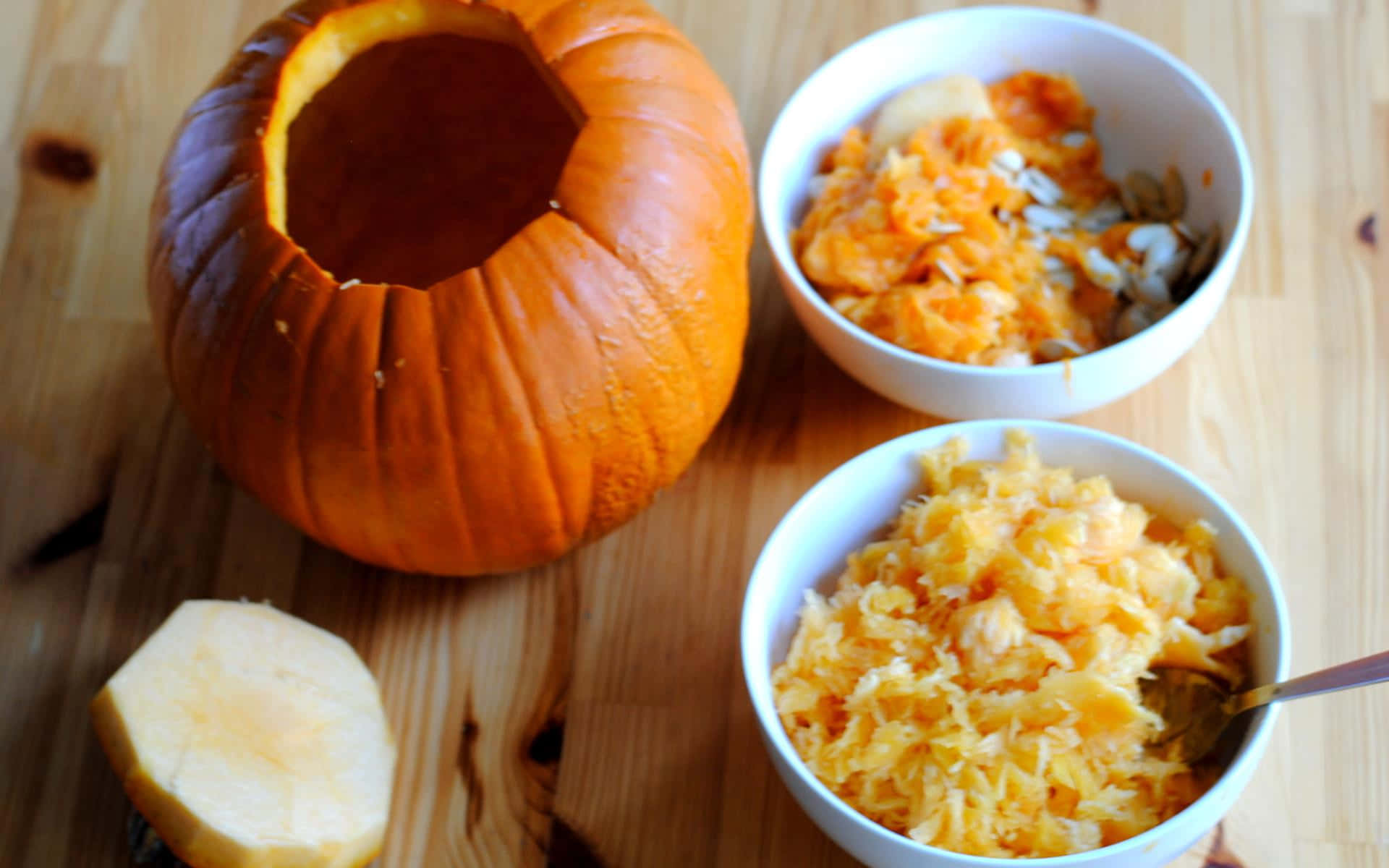 Autumn Food 桌布 壁紙 — 一個挖空的橙色南瓜放在溫暖的木桌上，旁邊有兩碗其亮橙色的果肉和種子。