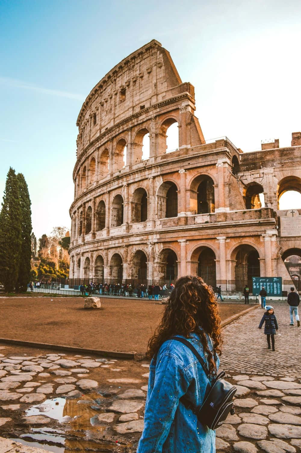 Rome 桌布 壁紙 — 一個捲髮、身穿藍色外套的人站在鵝卵石上，凝視著晴朗藍天下古老的羅馬競技場。