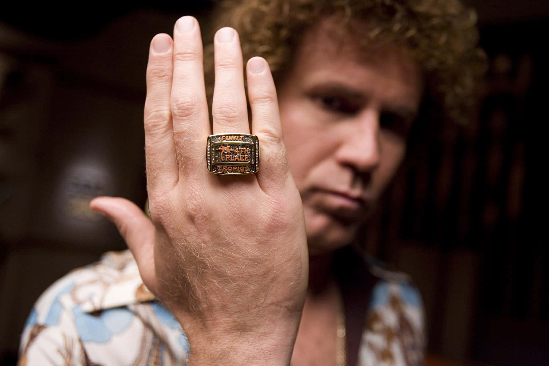 will ferrell 桌布 壁紙 — 一個捲髮男人舉起手，展示一隻刻有 'TROPICS' 的大型冠軍戒指。