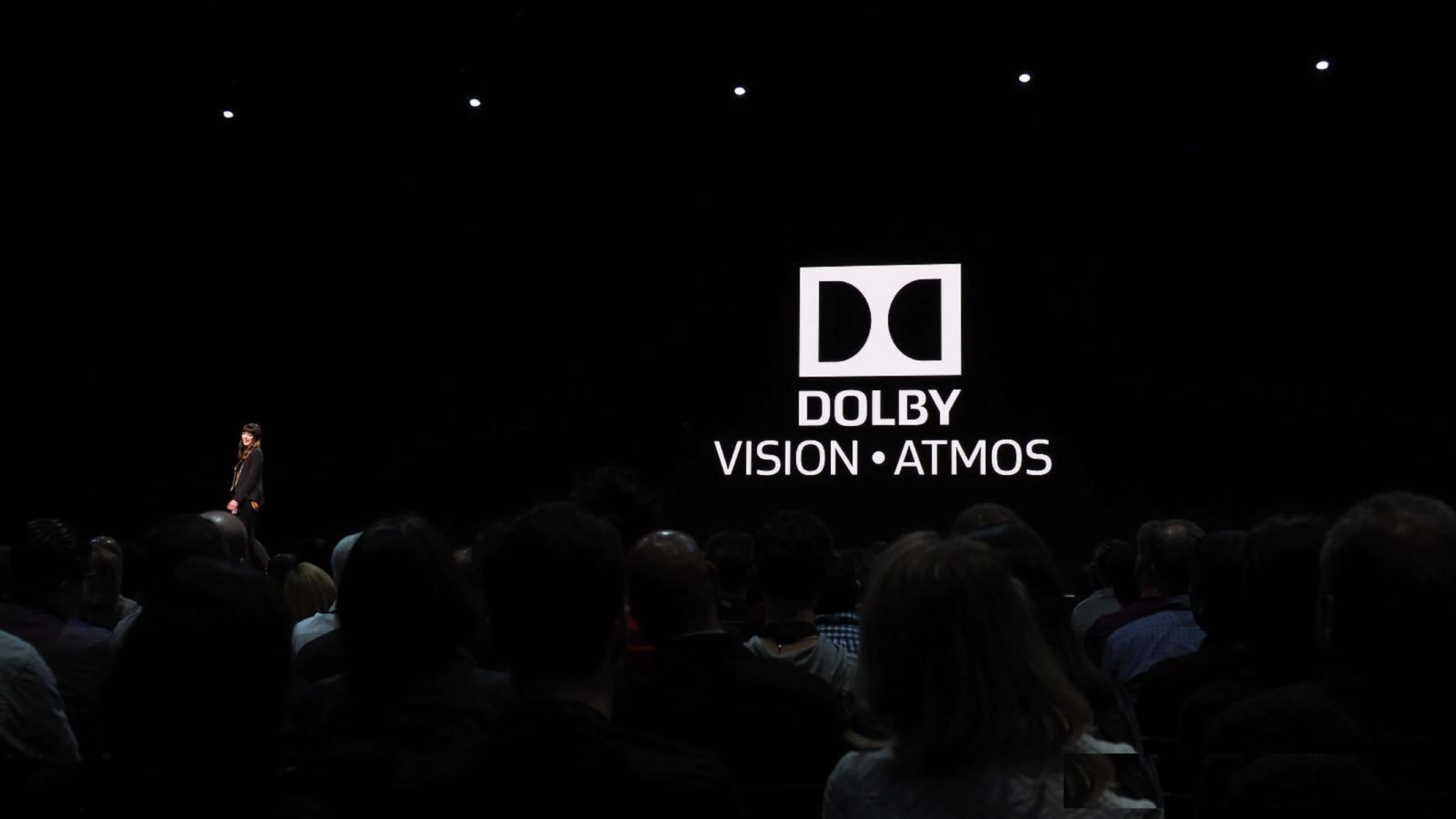 4K Dolby Vision 桌布 壁紙 — 一個揚聲器獨自站在黑暗的舞台上，面前是一個顯示Dolby Vision Atmos標誌的大屏幕，觀眾坐在昏暗的燈光中。