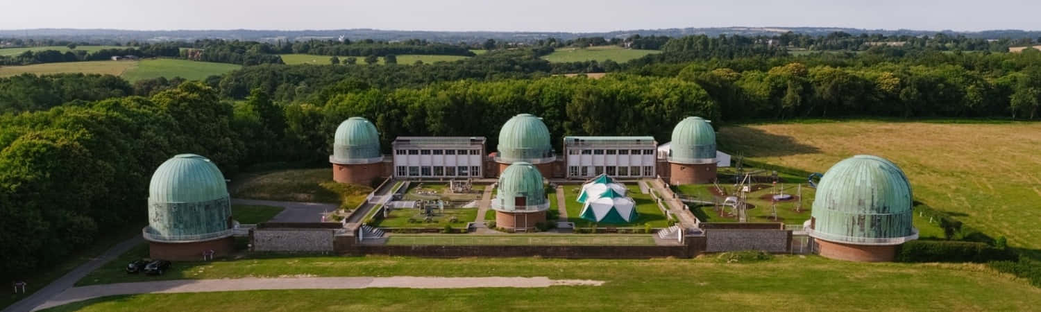 Observatory 桌布 壁紙 — 一個擁有五個綠色圓頂的宏偉天文台建築群的鳥瞰圖，坐落在晴朗天空下的鬱鬱蔥蔥的綠色景觀中。