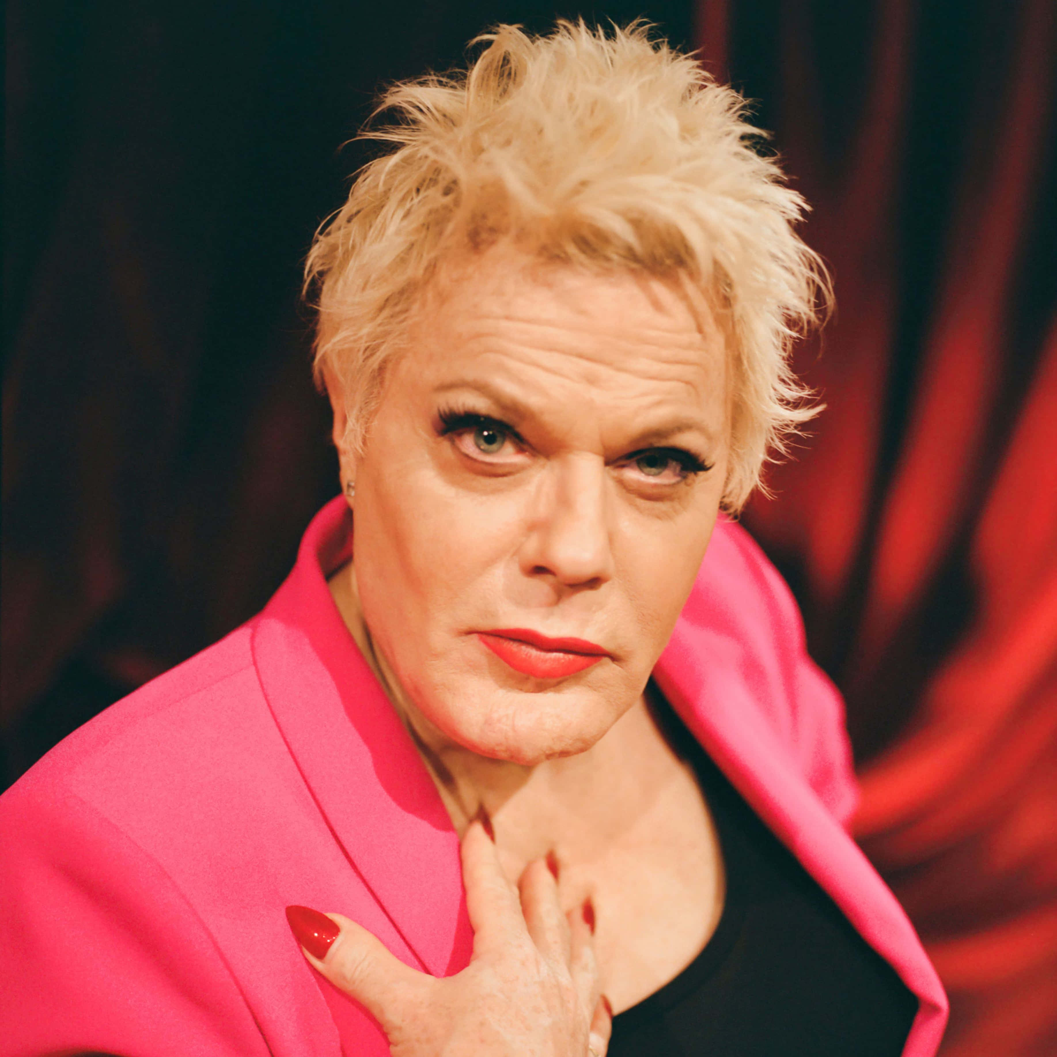 Eddie Izzard 桌布 壁紙 — 一個擁有尖刺金髮和鮮豔紅唇的人，穿著充滿活力的粉紅色西裝外套，在深紅色窗簾背景下自信地擺姿勢。