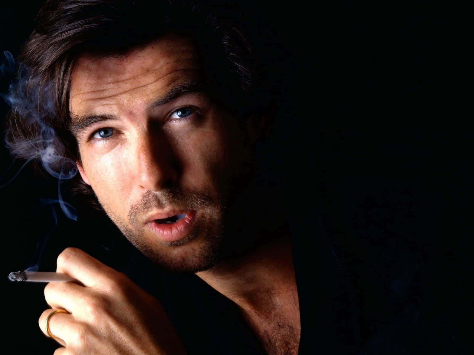 pierce brosnan 桌布 壁紙 — 一個擁有強烈藍眼的男人在黑暗背景下抽煙，煙霧向上盤旋。