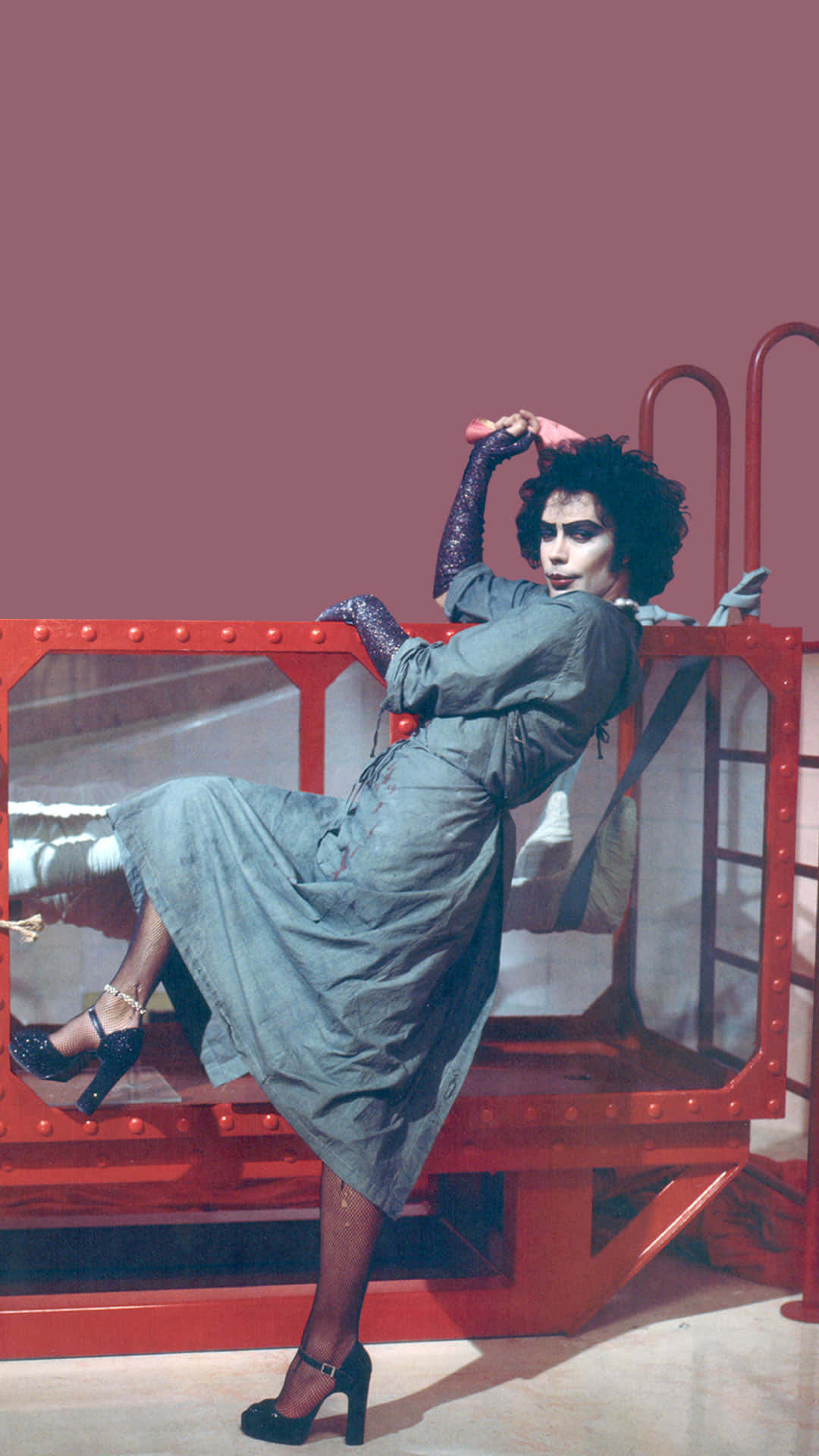 Tim Curry 桌布 壁紙 — 一個擁有捲髮和戲劇化妝容的人，在純色淡紫色背景上，於紅色工業結構上動態擺姿勢。