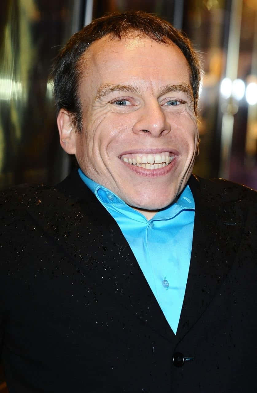 Warwick Davis 桌布 壁紙 — 一個擁有明亮藍眼睛和寬闊露齒笑容的男人，在鮮豔的藍色襯衫外穿著一件黑色閃亮外套。