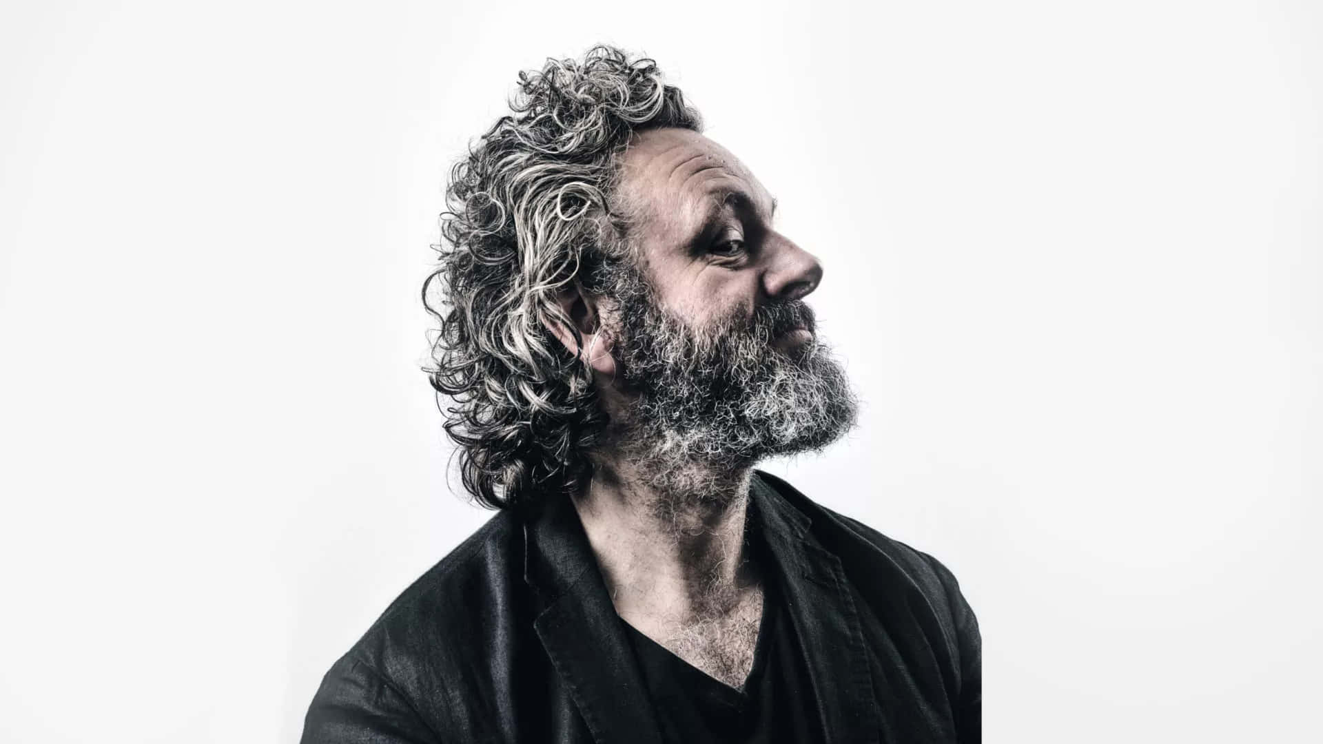 Michael Sheen 桌布 壁紙 — 一個擁有灰色捲髮和鬍鬚的男人，在純白背景上向上凝視，呈現高對比度的黑白肖像。