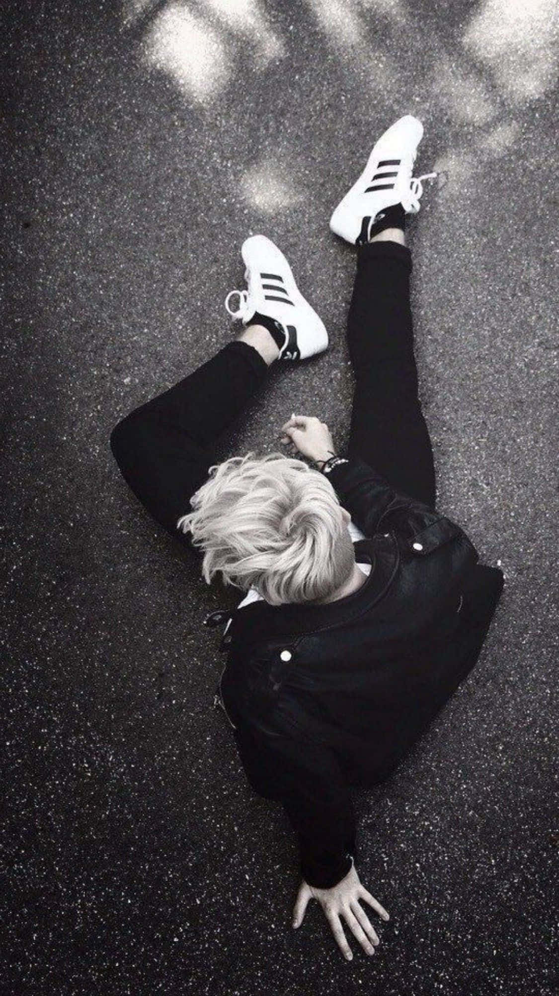 Bts Rap Monster 桌布 壁紙 — 一個擁有鉑金色頭髮的人躺在瀝青路上，穿著黑色衣服和白色運動鞋，在斑駁的陽光下。