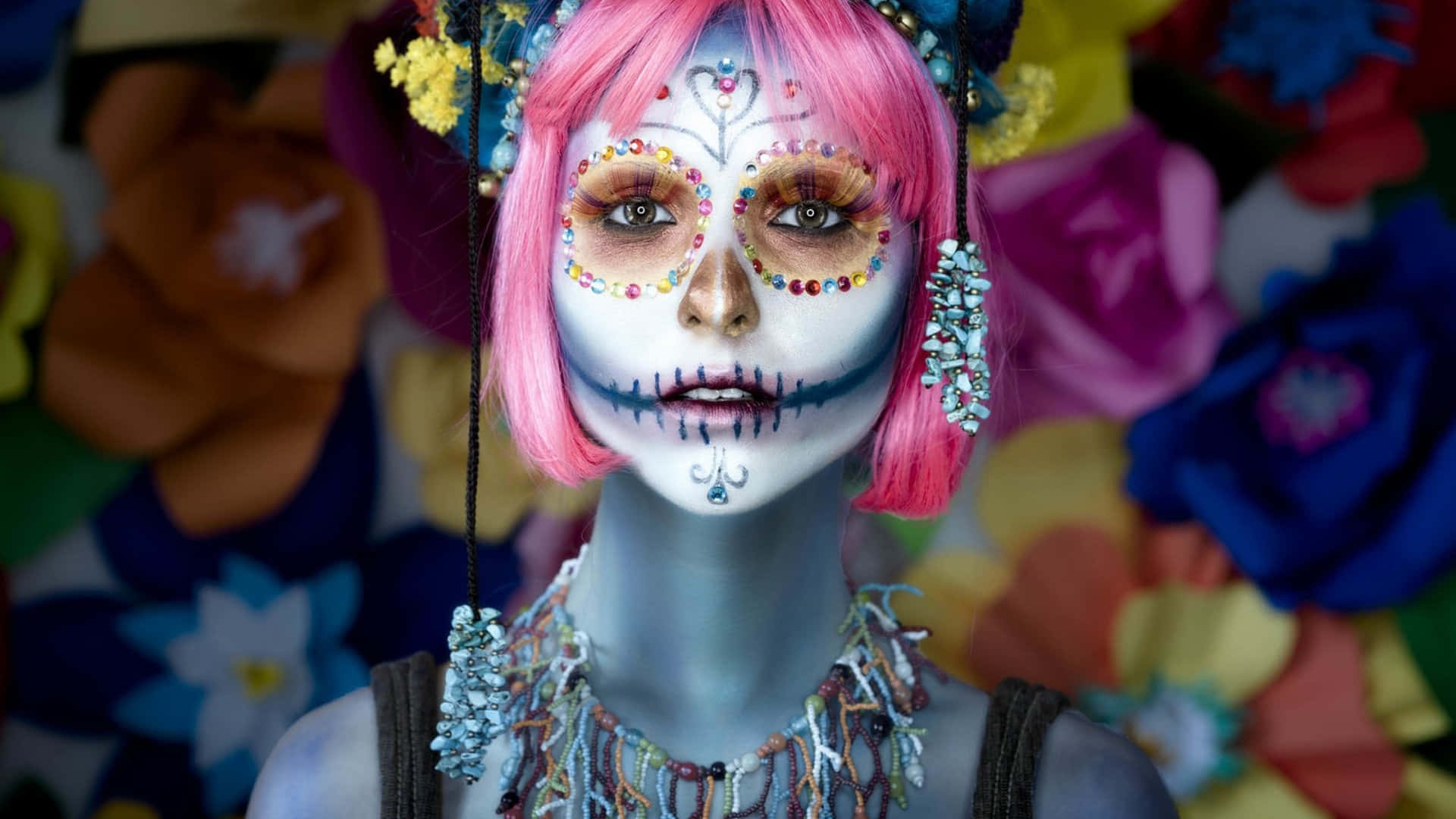 Halloween Makeup 桌布 壁紙 — 一個擁有鮮豔粉紅色頭髮和精緻糖骷髏妝容的人，站在色彩繽紛的大型紙花背景前。