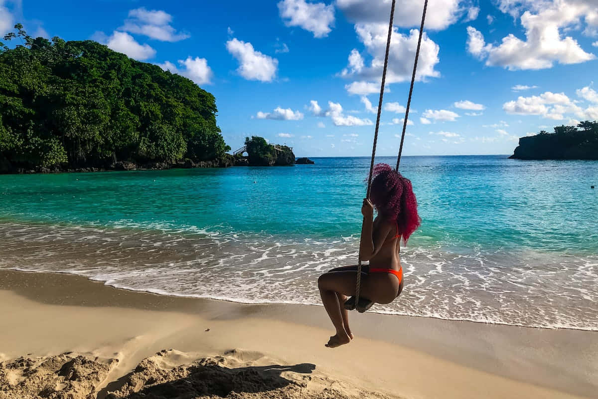 Jamaican Island 桌布 壁紙 — 一個擁有鮮豔紅髮的人喺陽光普照嘅沙灘上，用繩索鞦韆喺碧綠嘅海水同茂密嘅綠色懸崖下，喺明亮嘅藍天下搖擺。