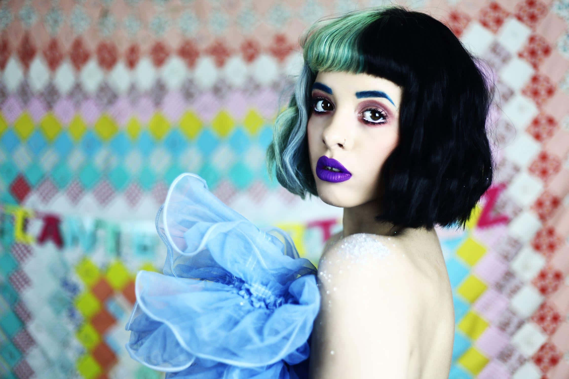 Melanie Martinez 桌布 壁紙 — 一個擁有鮮豔紫色嘴唇和藍黑色頭髮的人，在色彩繽紛的拼布被子背景前擺姿勢。