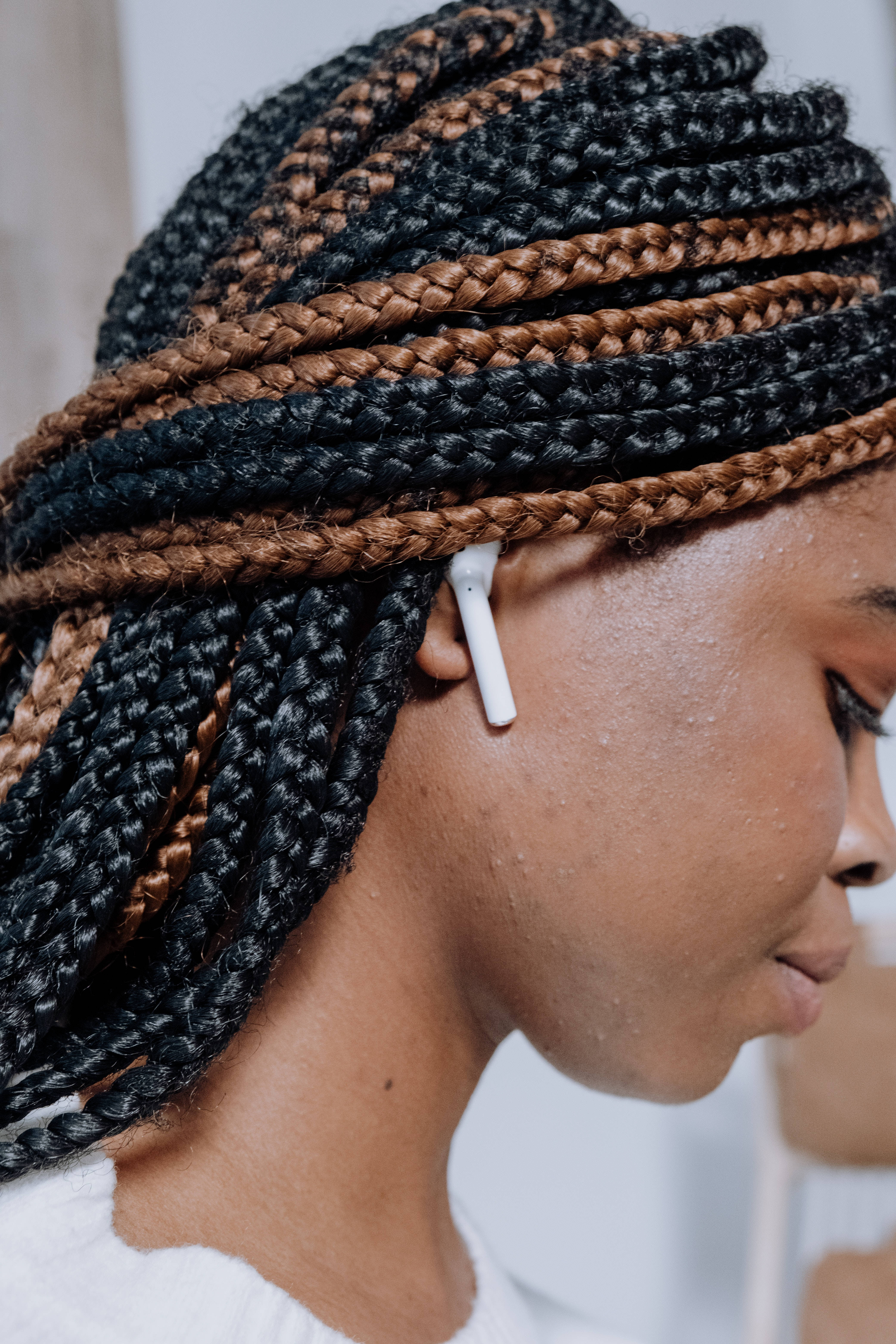 Airpods 桌布 壁紙 — 一個擁有黑色和棕色辮子頭髮的人，側面佩戴白色無線耳塞的特寫。