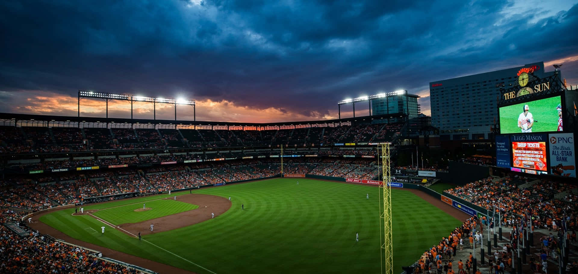 MLB Baseball League 桌布 壁紙 — 一個擠滿球迷的棒球場，在戲劇性的黃昏天空下，有橙色雲朵和發光的球場燈光。