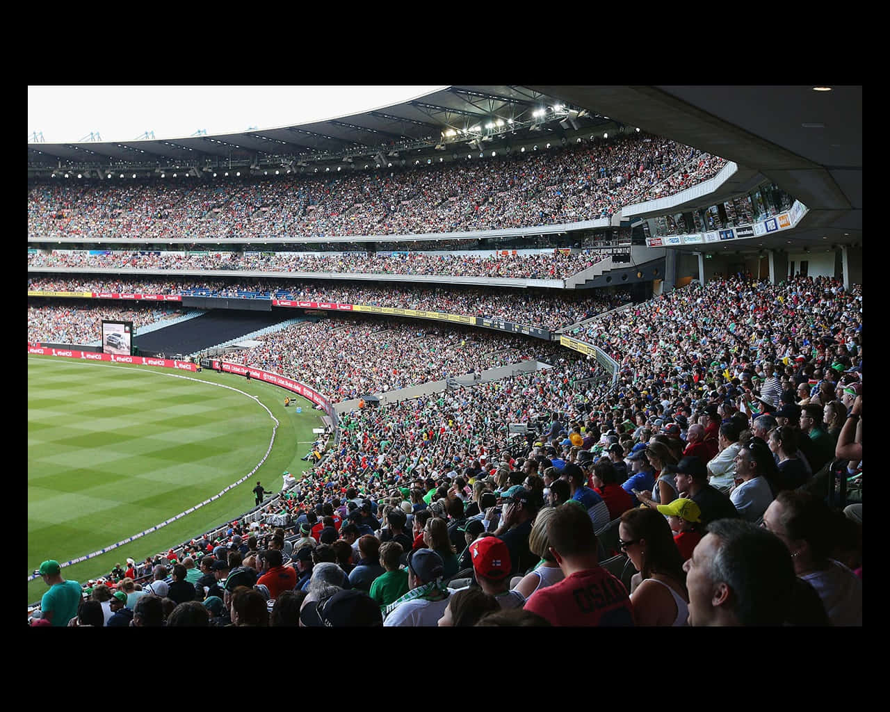 Cricket Ground 桌布 壁紙 — 一個擠滿球迷的體育場，球迷們穿著色彩繽紛的服裝，在明亮的日光下圍繞著 pristine 的綠色板球場。