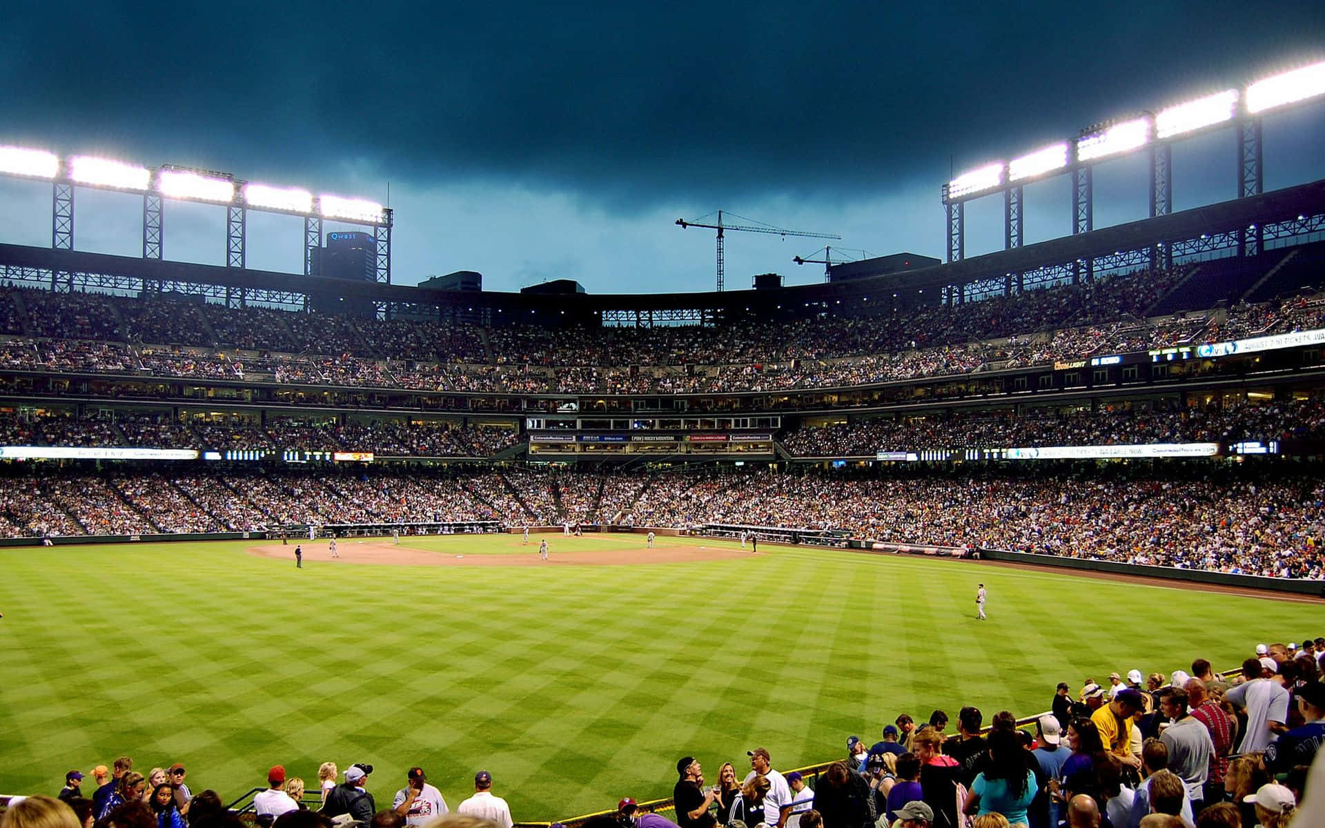 Baseball Stadium 桌布 壁紙 — 一個擠滿觀眾的棒球場，在戲劇性的夜空下，明亮的泛光燈照亮了綠色球場和歡呼的人群。