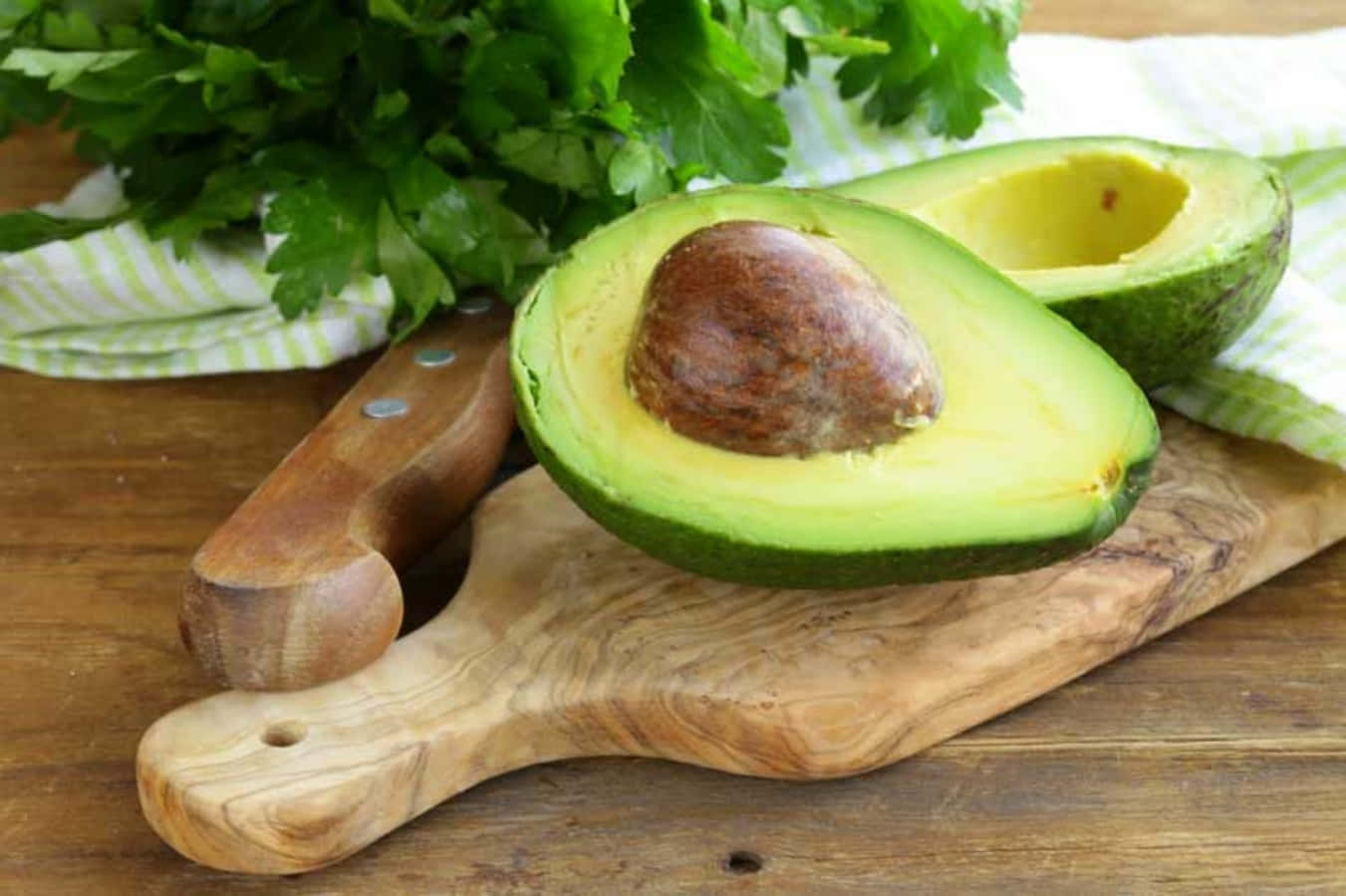 Avocado 桌布 壁紙 — 一個新鮮切開的牛油果，帶有一個大的棕色果核，放在木砧板上，旁邊有一把刀和歐芹。