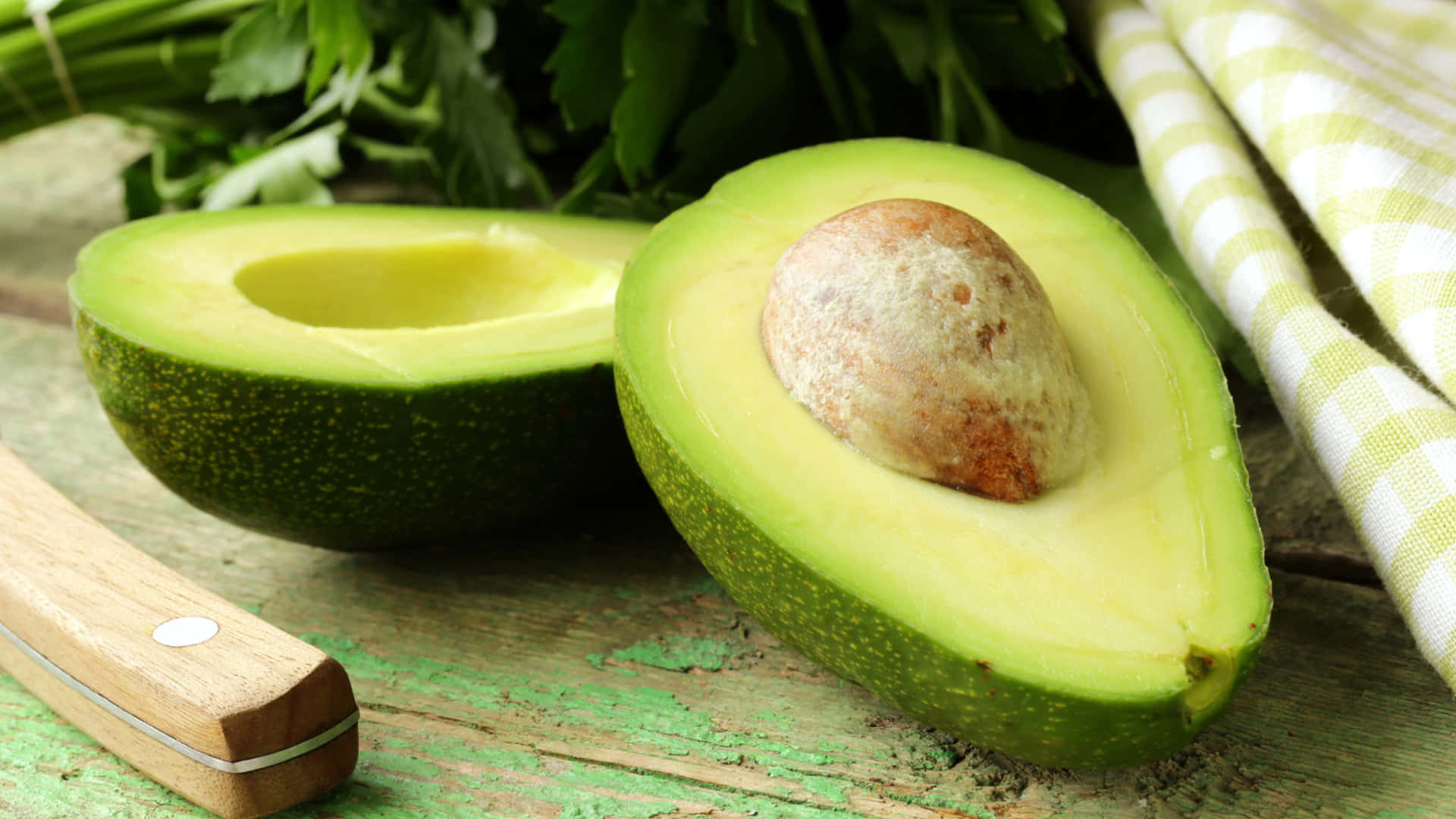Avocado 桌布 壁紙 — 一個新鮮切開的牛油果，帶有大的棕色果核，放在一個風化的綠色木質表面上，旁邊有歐芹和一塊格子布。