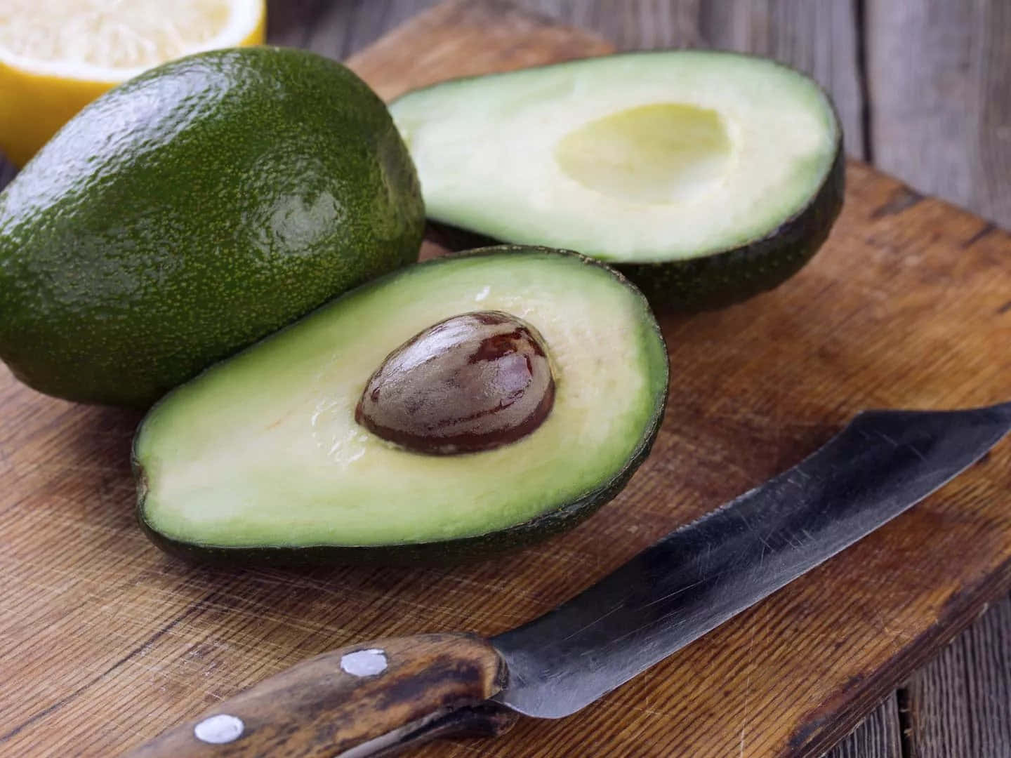 Avocado 桌布 壁紙 — 一個新鮮牛油果切半，果核外露，放在一個質樸的木砧板上，旁邊有一把刀和一片檸檬。