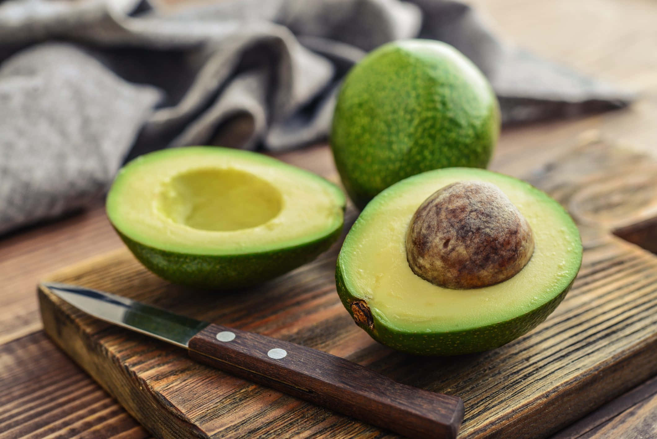 Avocado 桌布 壁紙 — 一個新鮮牛油果切半放在質樸的木砧板上，旁邊有一把刀。