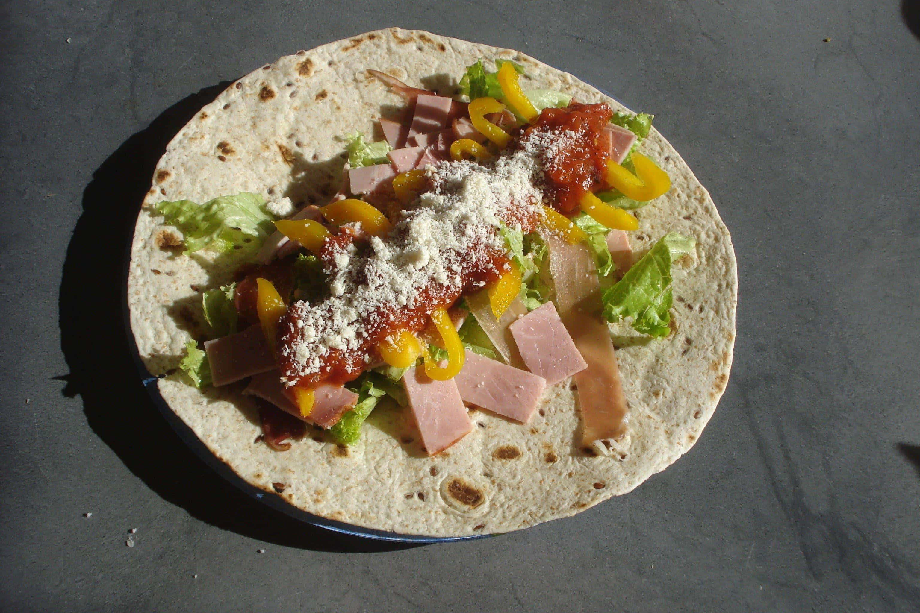 Taco 桌布 壁紙 — 一個新鮮組裝的玉米餅，裡面有火腿、黃椒、生菜和芝士，放在深色碟子上，在明亮的陽光下。