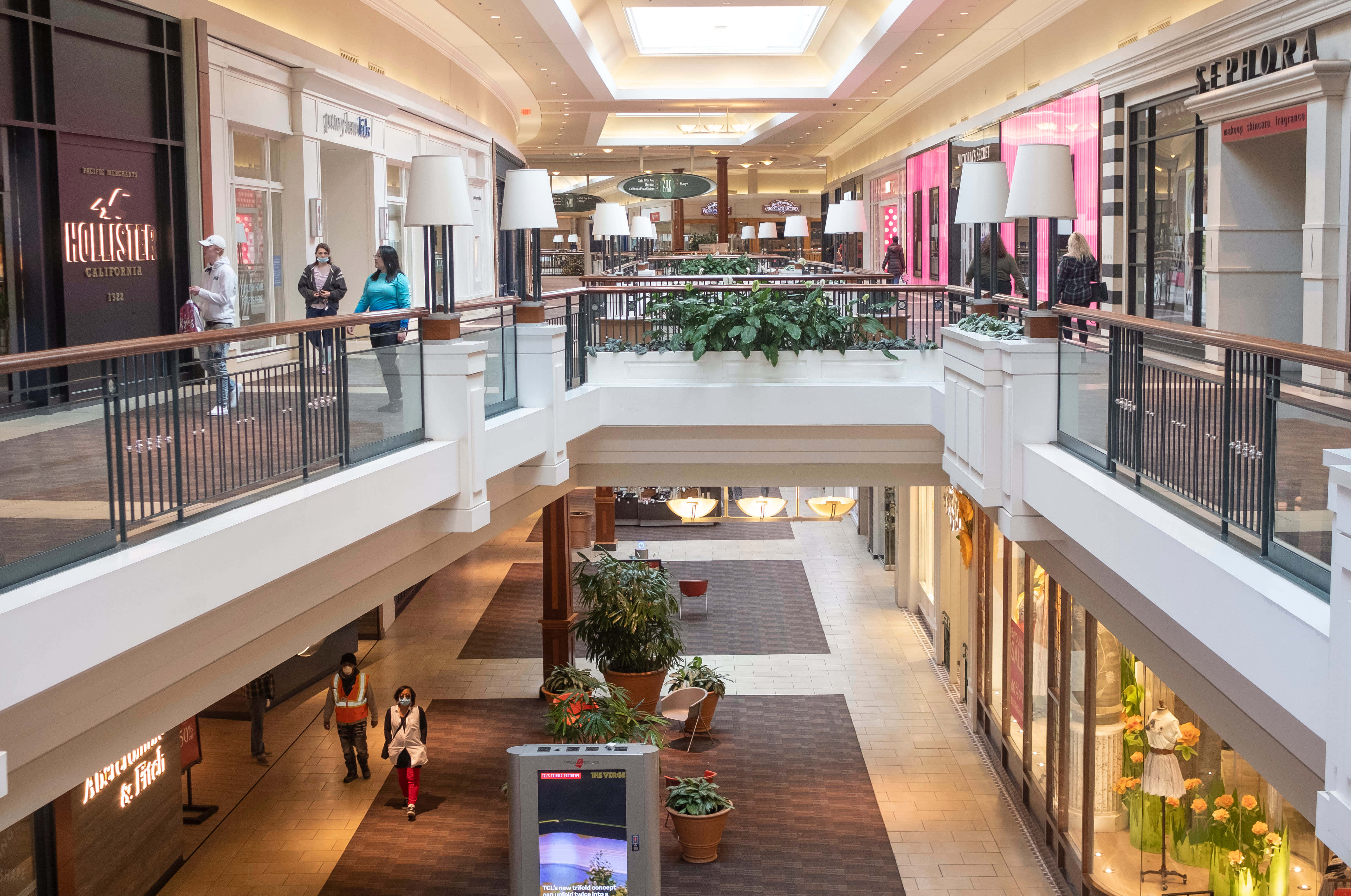 Mall 桌布 壁紙 — 一個明亮的多層購物中心，購物者在發光的天花板燈光下走過 Hollister 和 Sephora 等商店。