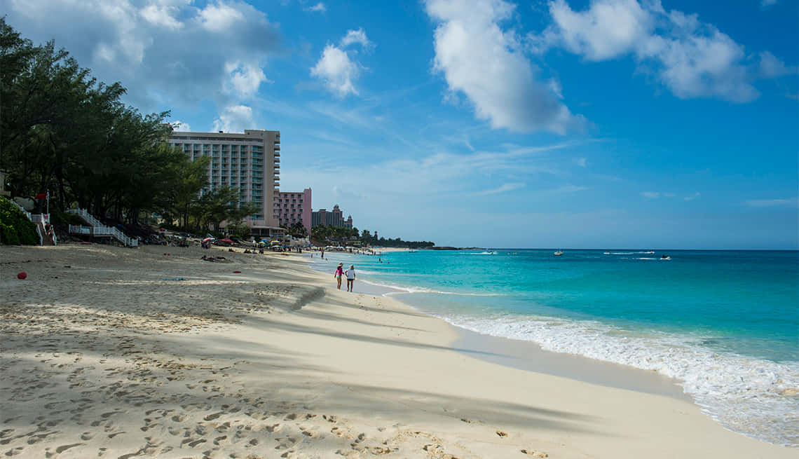 Bahamas Island 桌布 壁紙 — 一個明亮的陽光沙灘，有碧綠的海水、溫柔的海浪和一個大型酒店綜合體，在藍天白雲下。