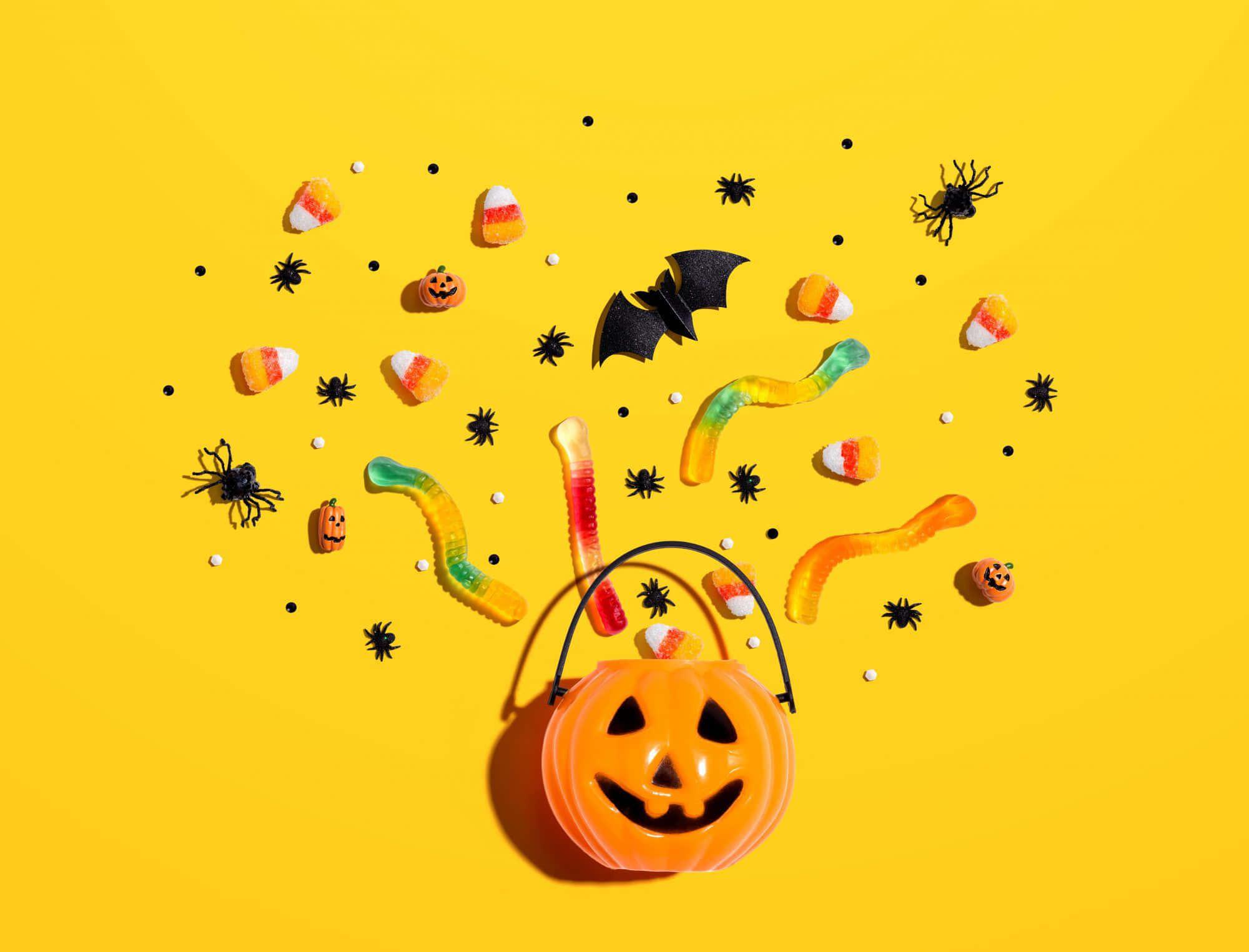Halloween Treats 桌布 壁紙 — 一個明亮的黃色背景，散落著萬聖節糖果、軟糖蟲、糖果玉米、蜘蛛和一個微笑的南瓜桶。
