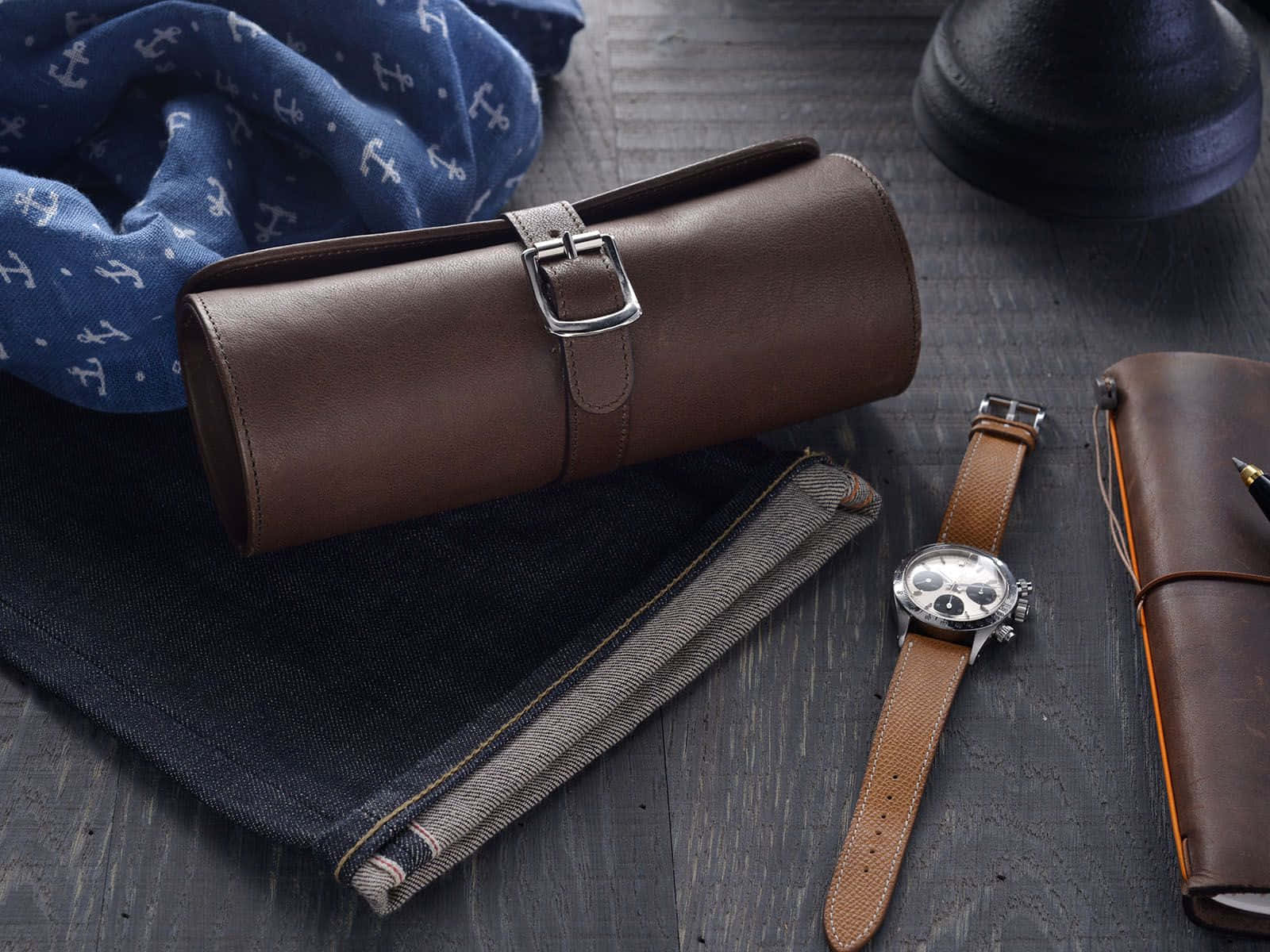 Accessory 桌布 壁紙 — 一個時尚的平鋪擺拍，包括棕色皮革手錶盒、計時碼錶、牛仔褲和藍色船錨圍巾，放置在深色木頭上。